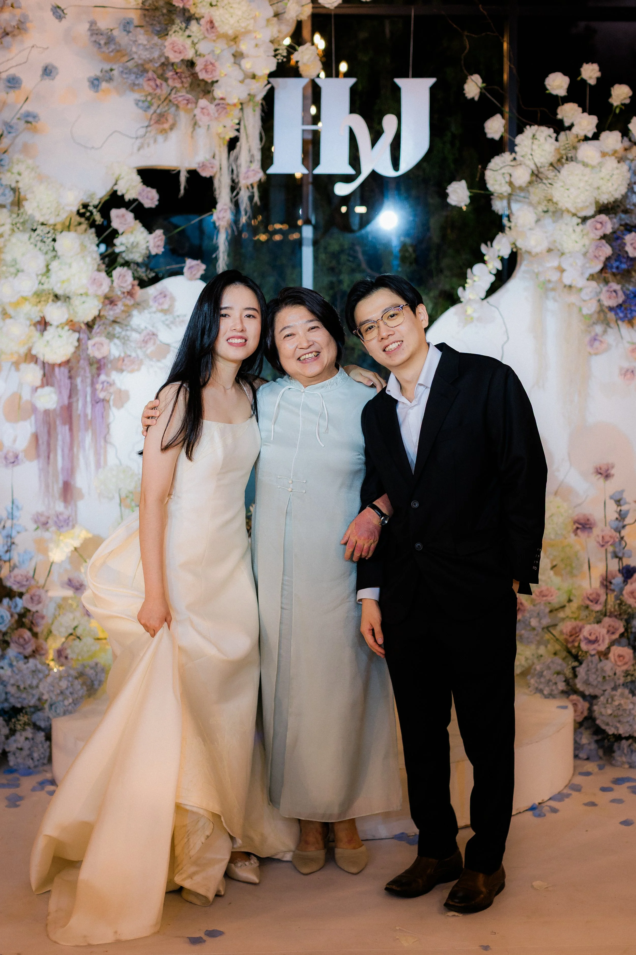 Three people, a woman in a cream-colored dress, an older woman in a light blue dress, and a man in a black suit, are standing close together and smiling at a floral decorated party with a sign that says "Hi" above them.