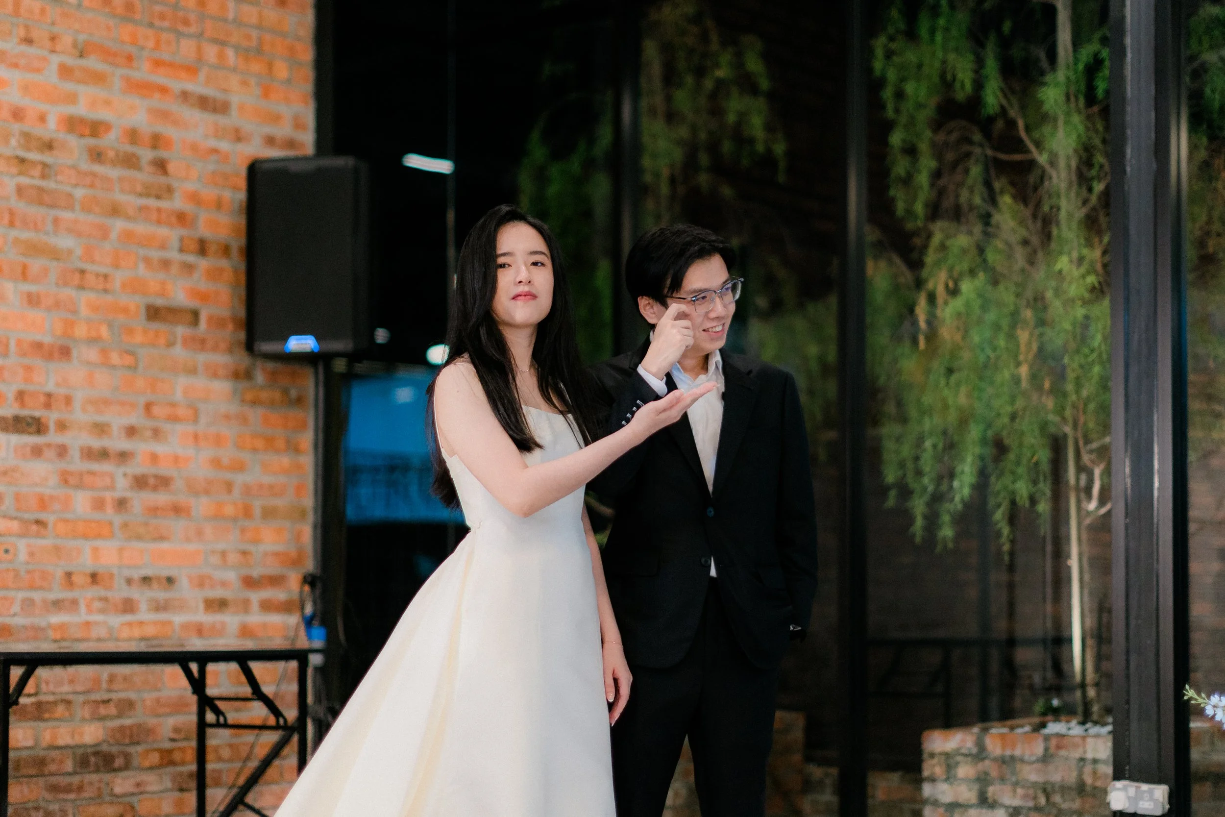 A young woman in a white dress and a young man in a black suit and glasses standing indoors near a large window with greenery outside, with the woman making a gesture as if guiding or explaining something.