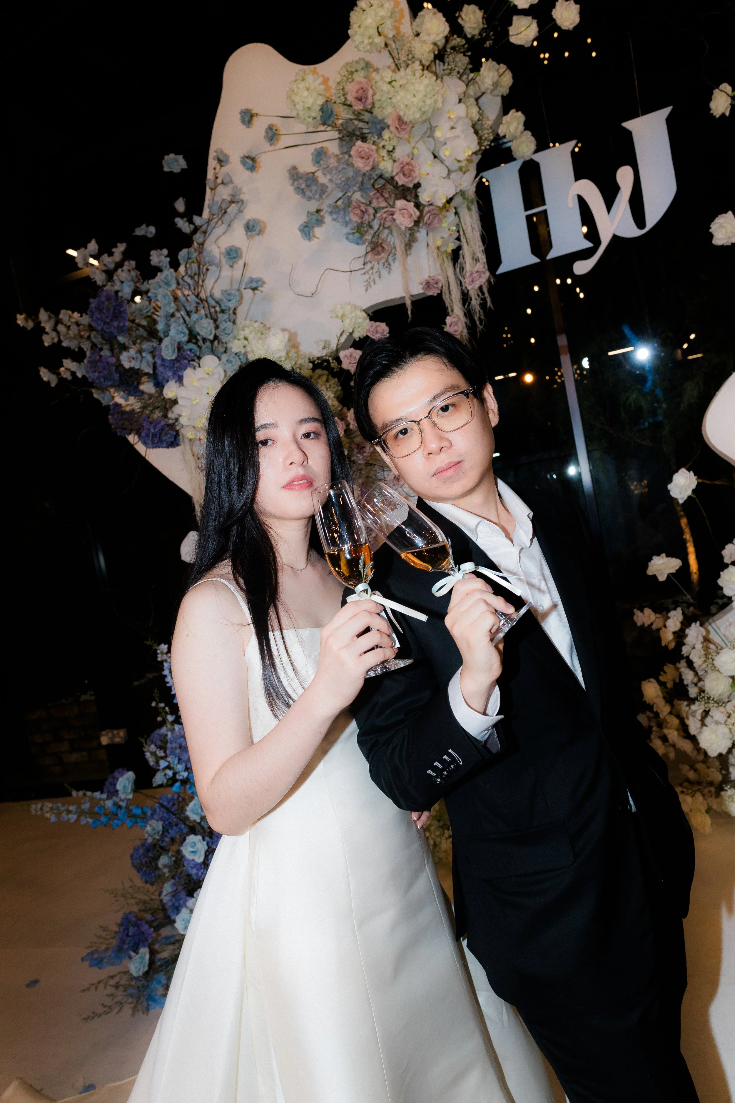 A young man and woman in formal attire holding glasses of champagne at a celebration, with floral decorations and the word 'HAPPY' in the background.