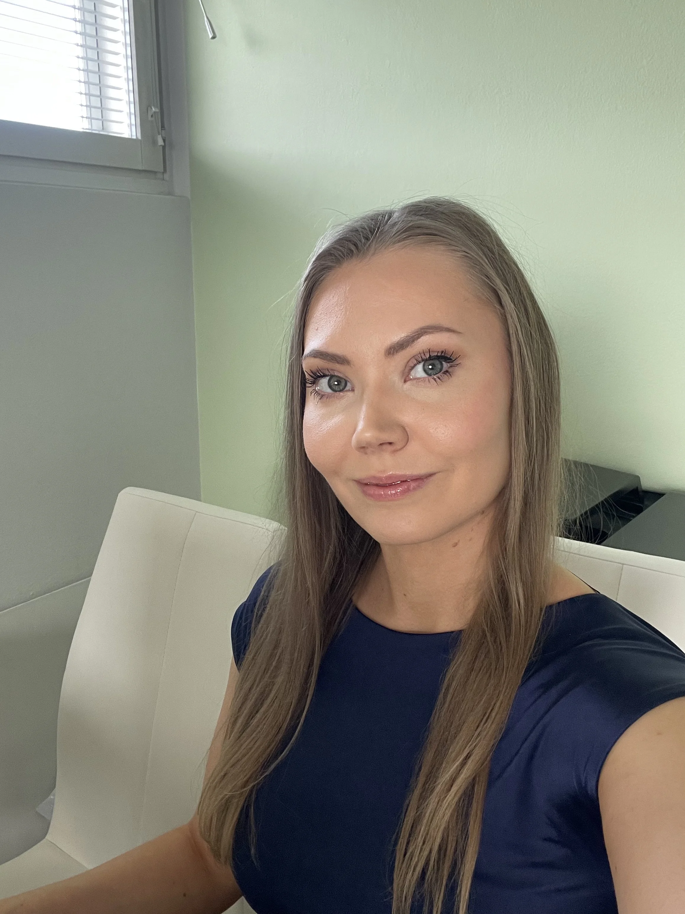 A woman with long, straight, light brown hair and striking blue eyes is taking a selfie indoors. She is wearing a dark blue top and is smiling softly. The background features a light green wall, a window with blinds, and a white chair.