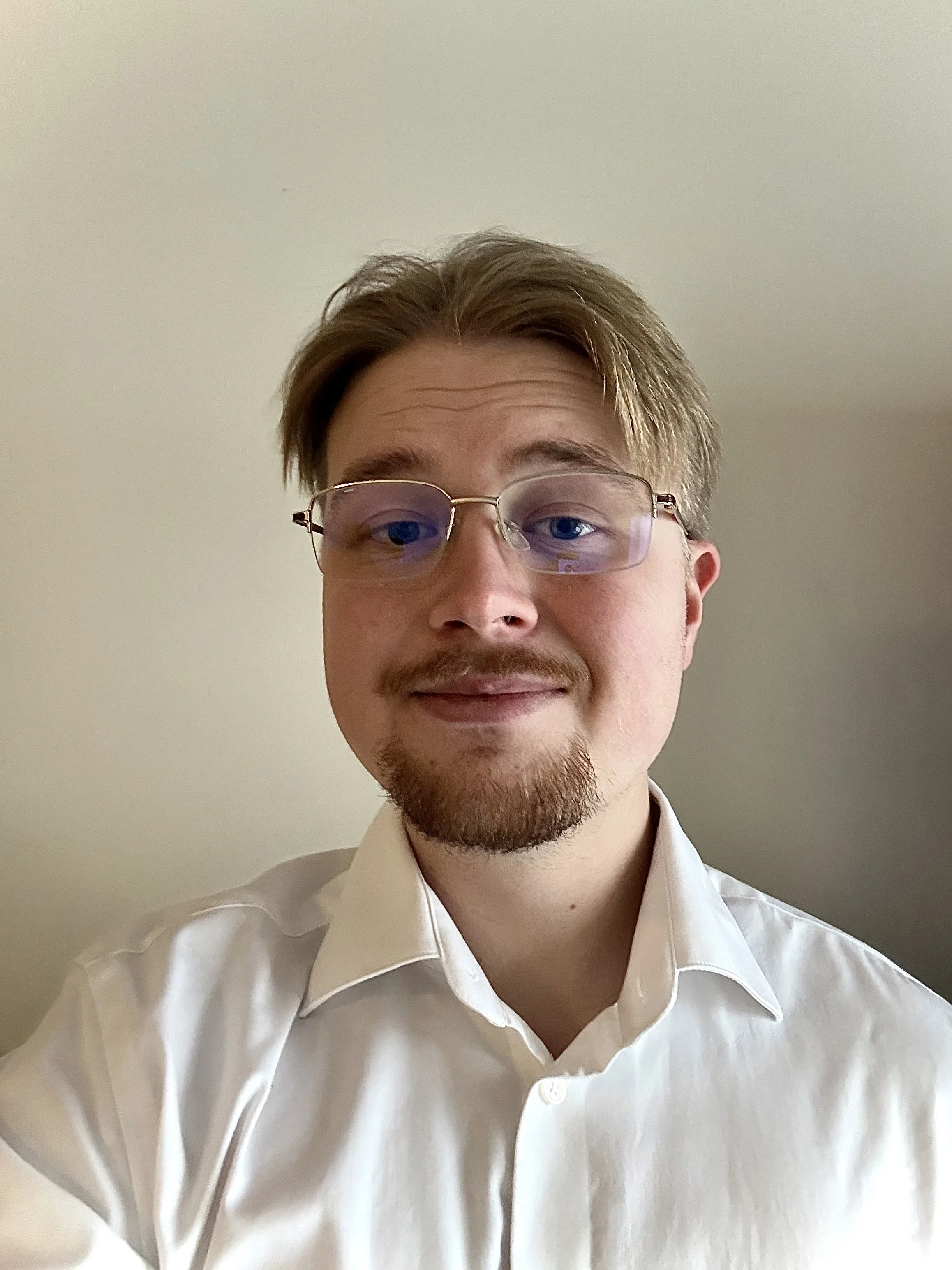 A young man with light skin, brown hair, glasses, and a goatee, wearing a white dress shirt, smiling slightly, in front of a plain beige wall.