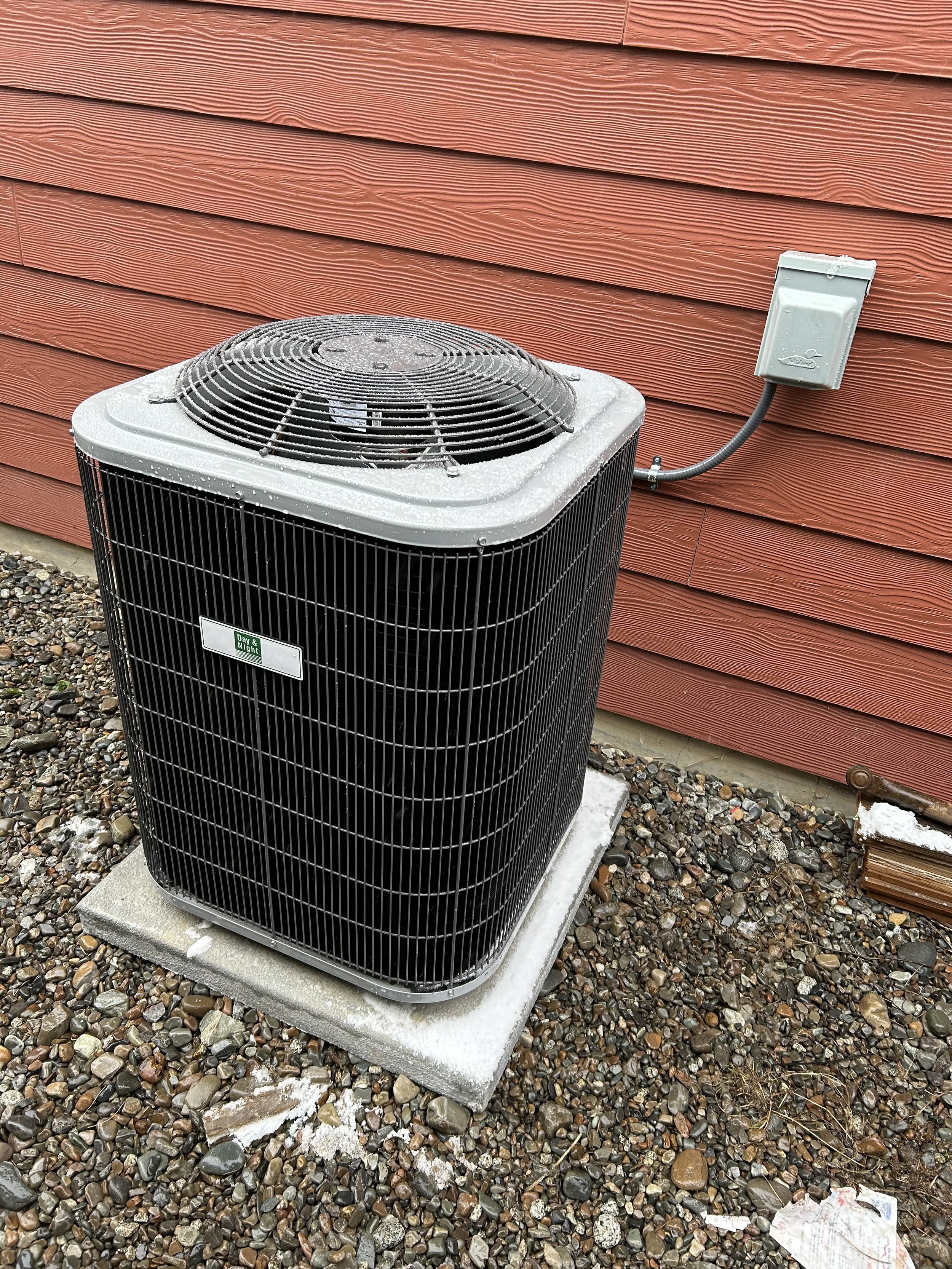 Outdoor air conditioning unit installed next to a red wooden house, sitting on a concrete pad, with frost or ice on the top and surrounding gravel ground.
