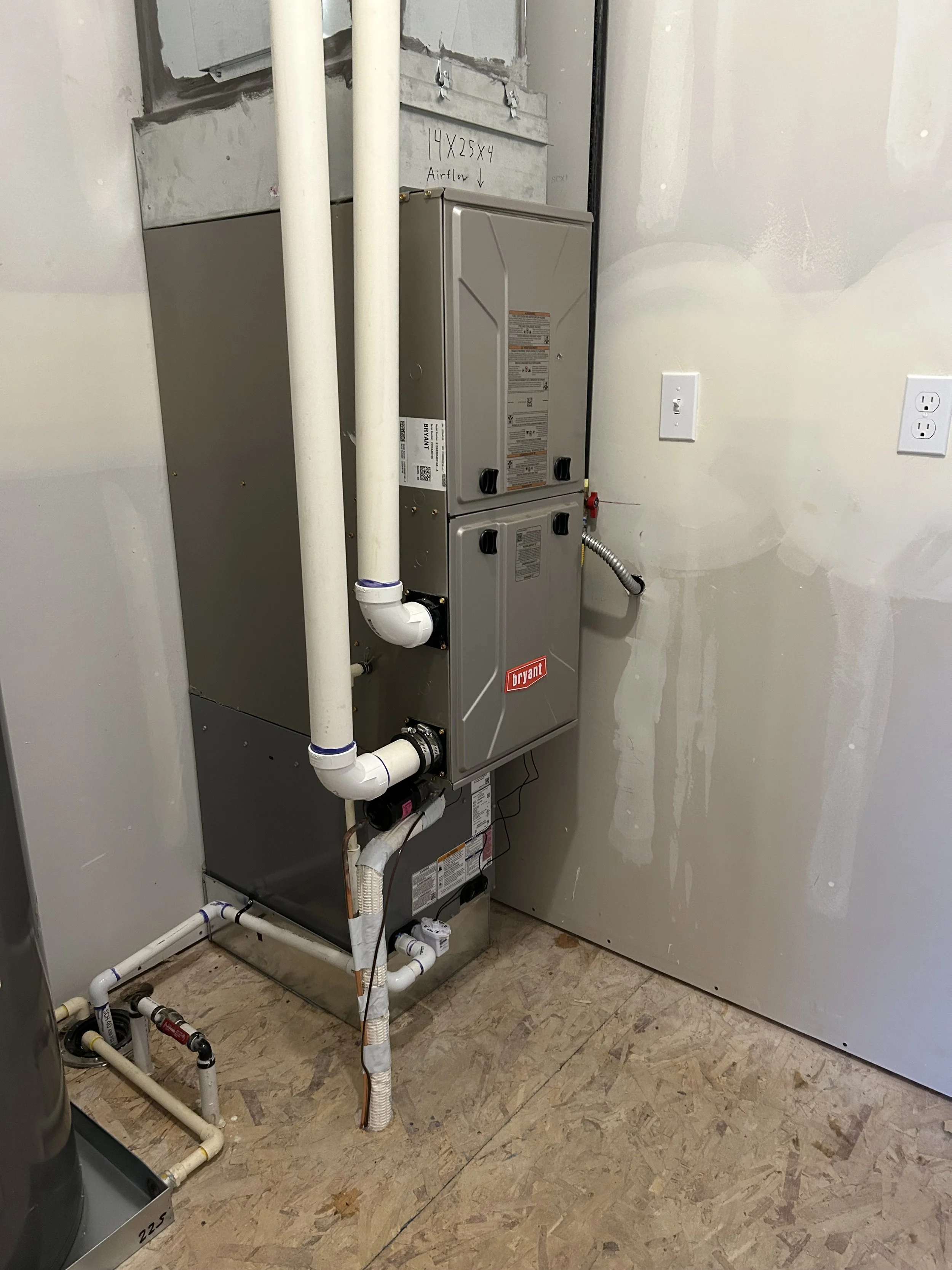 A Bryant HVAC unit with white PVC pipes connected, installed in a partially finished room with drywall and electrical outlets visible.