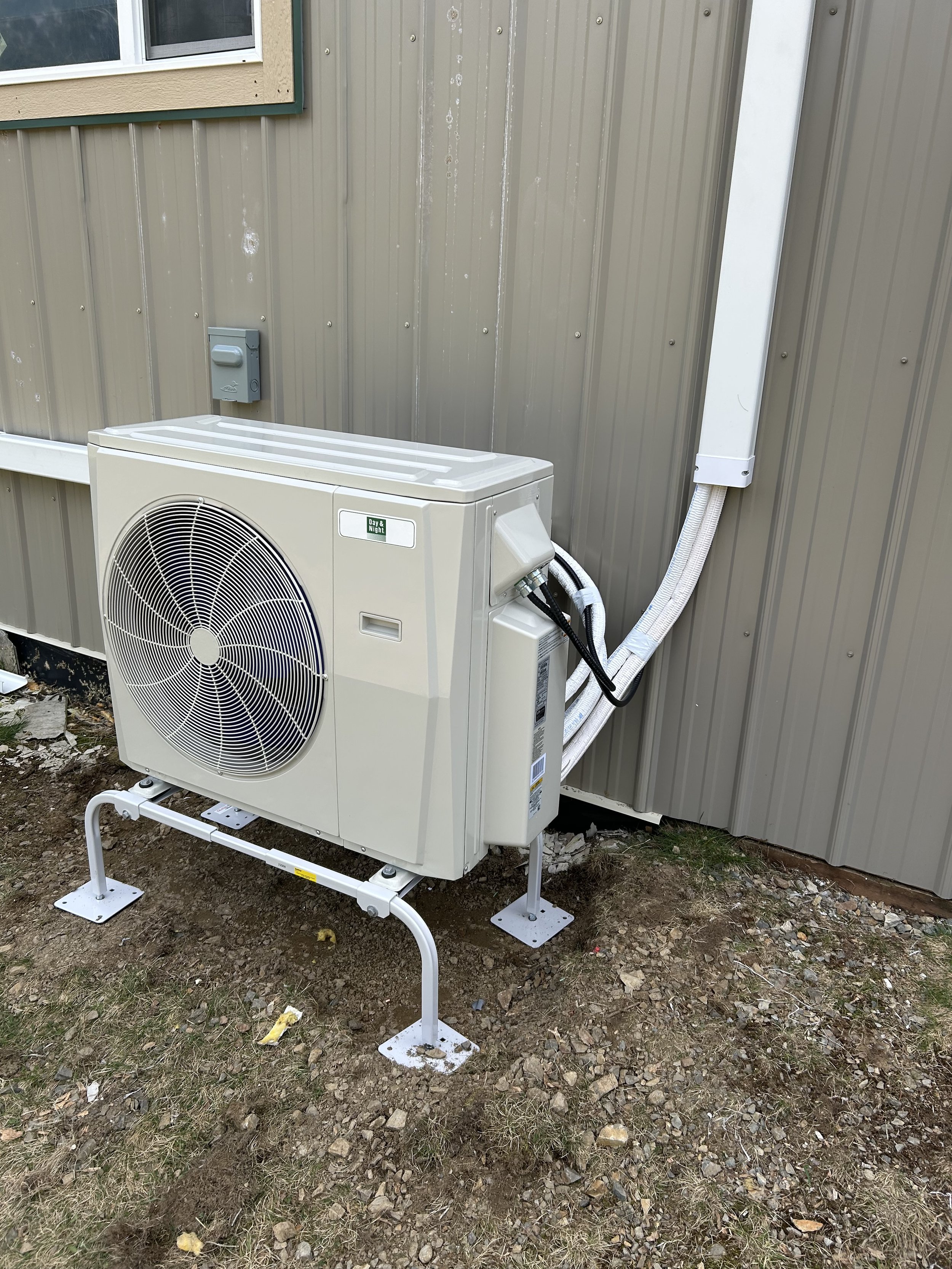 Outdoor HVAC compressor unit installed next to a beige metal building wall, elevated on a metal frame with screws into the ground, with electrical connections and conduit attached.