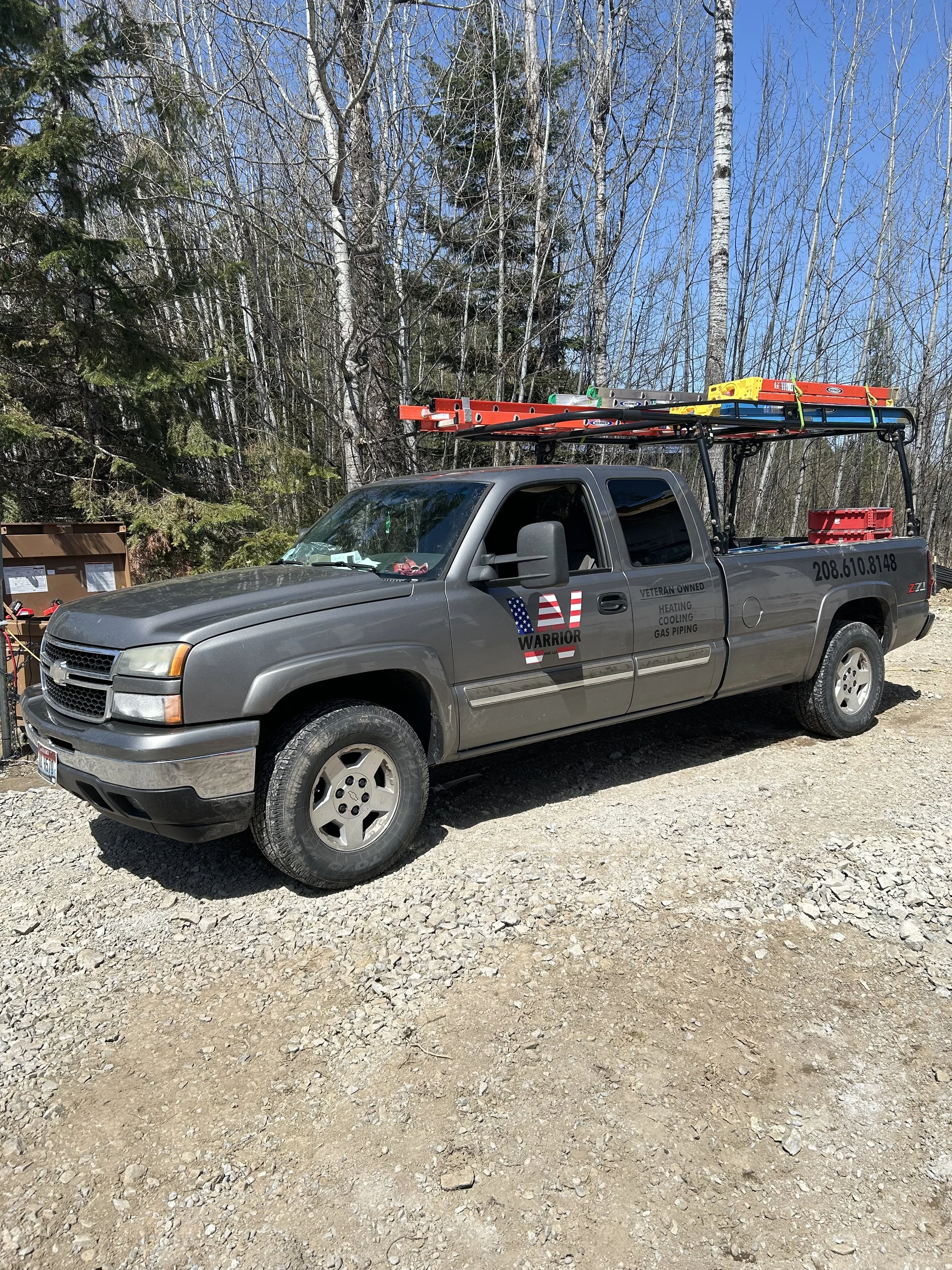 Grey pickup truck parked outdoors, with ladders and equipment on the roof. The truck has a logo on the side that says "Warrior" with an American flag design. The background has trees with no leaves, indicating early spring or late fall.