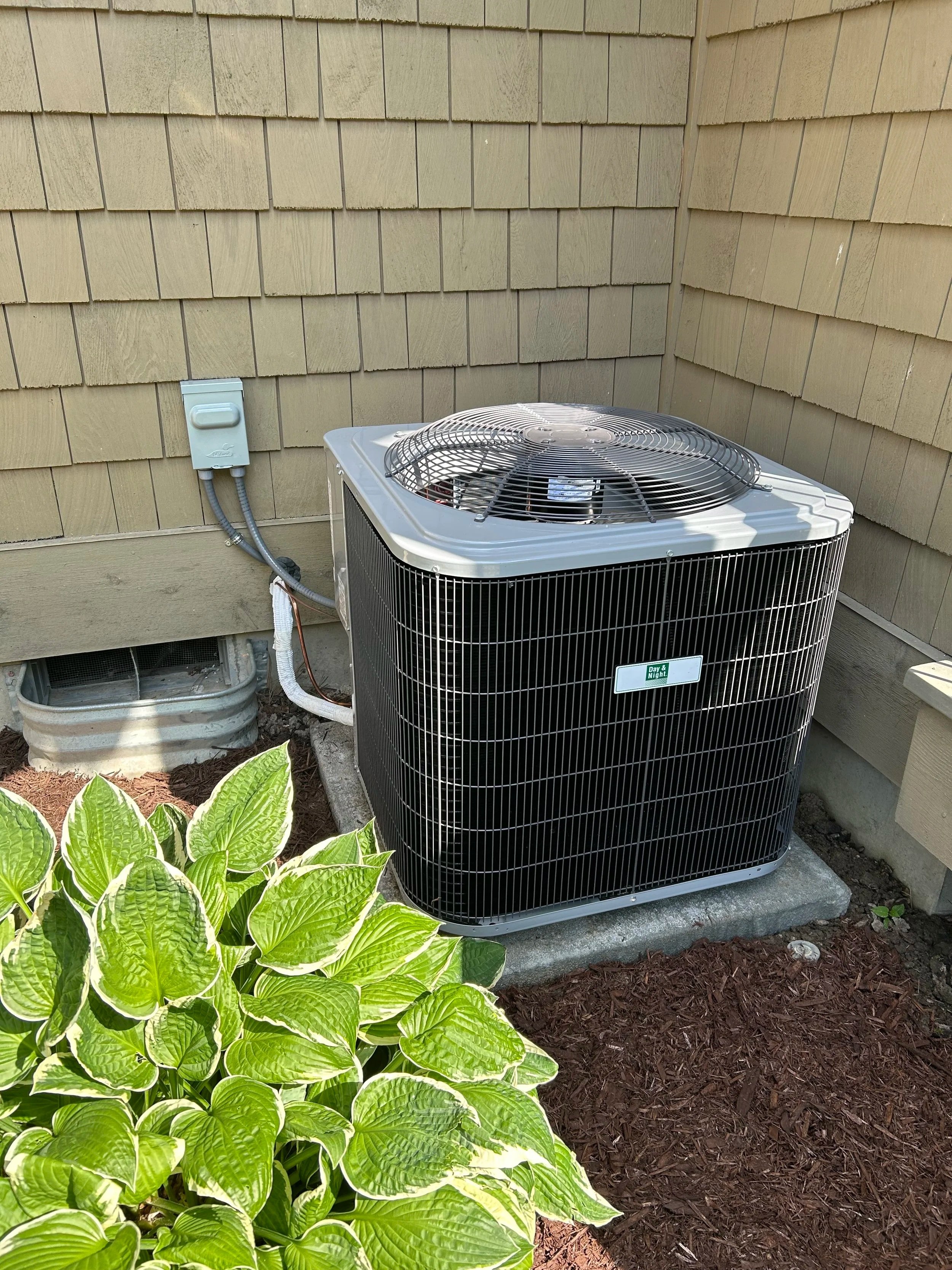 An outdoor air conditioning unit next to a house with beige siding, surrounded by some green leafy plants and brown mulch.