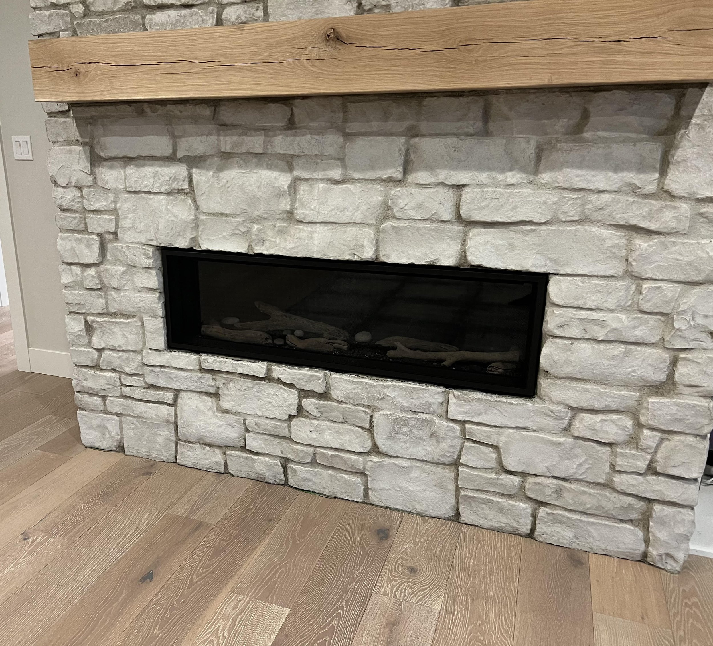 A white stone fireplace with a rectangular glass insert and a wooden mantel above it, located on a hardwood floor.