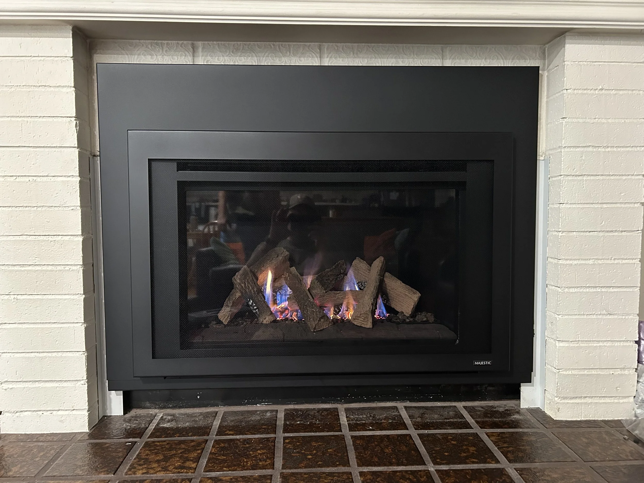 Electric fireplace with logs, flames, and purple and blue fire effects, surrounded by white brick wall and brown tiled hearth.