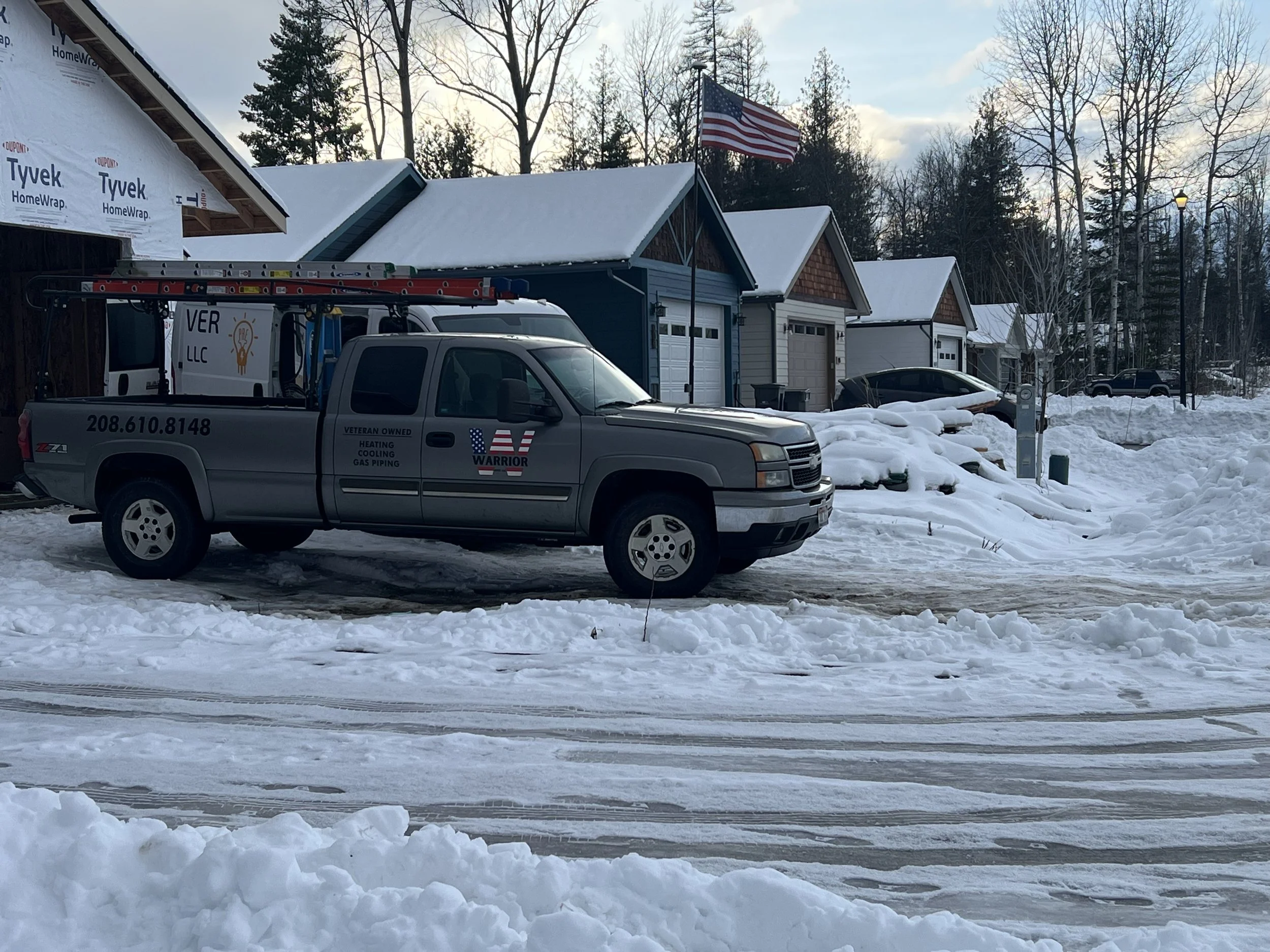 A snow-covered residential street with houses and parked cars. A gray utility truck with ladder on top, labeled with plumbing and heating service, is parked in the foreground. An American flag is flying in the background.