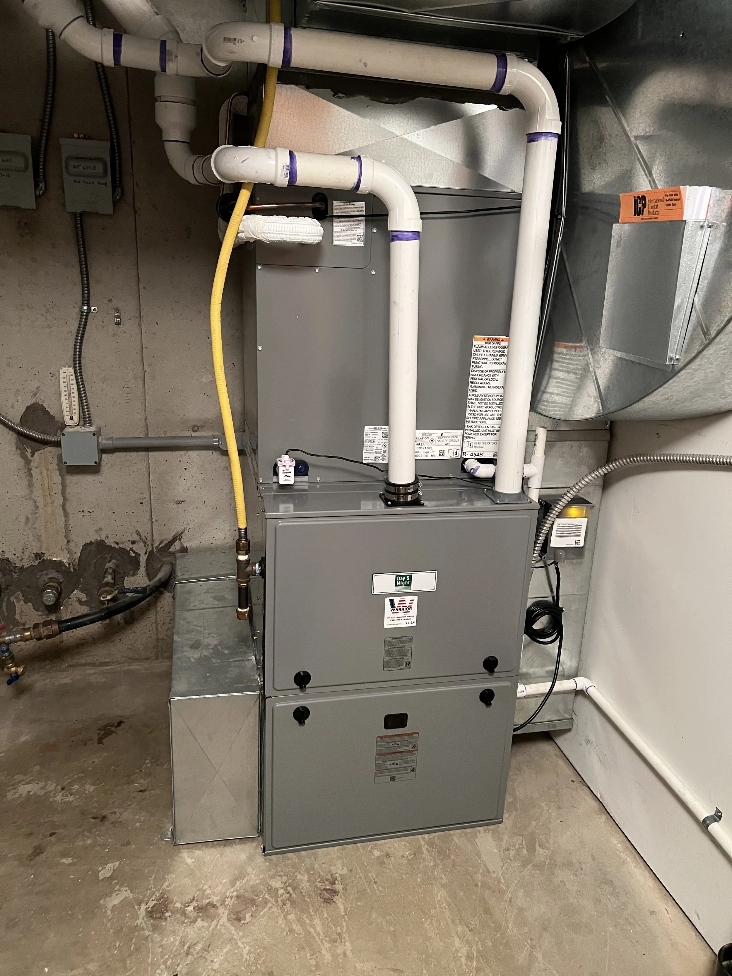 An HVAC furnace installed in a utility room with white pipes, a metallic vent, electrical wiring, and a concrete floor.