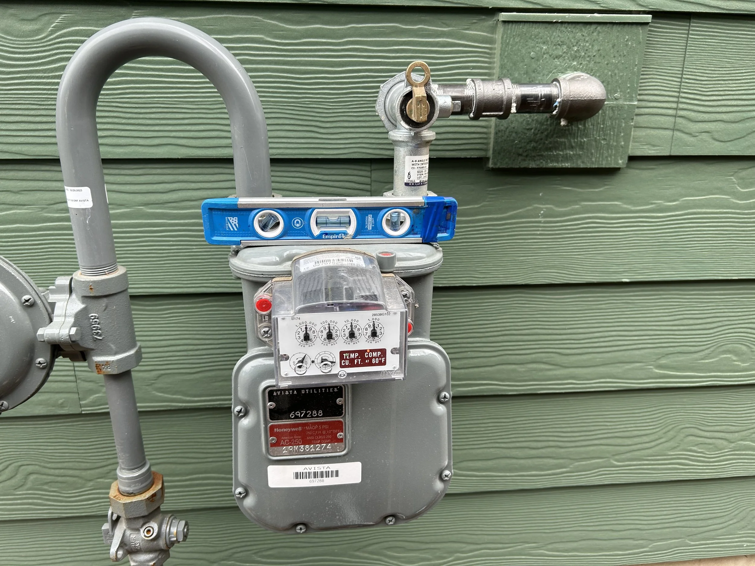 An outdoor utility gas meter with pipes attached, installed against a green wooden wall.