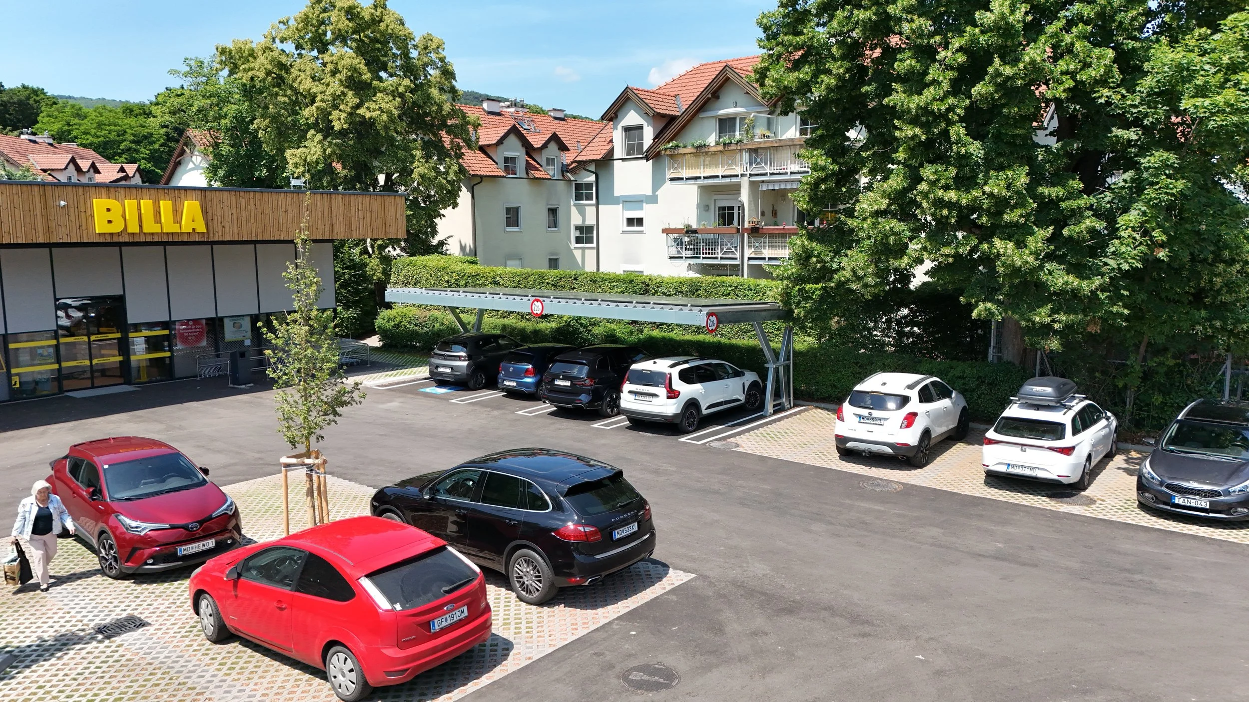 Ein Parkplatz neben einem Supermarkt namens 'BILLA'. Es gibt mehrere parkende Autos und Bäume im Hintergrund.
