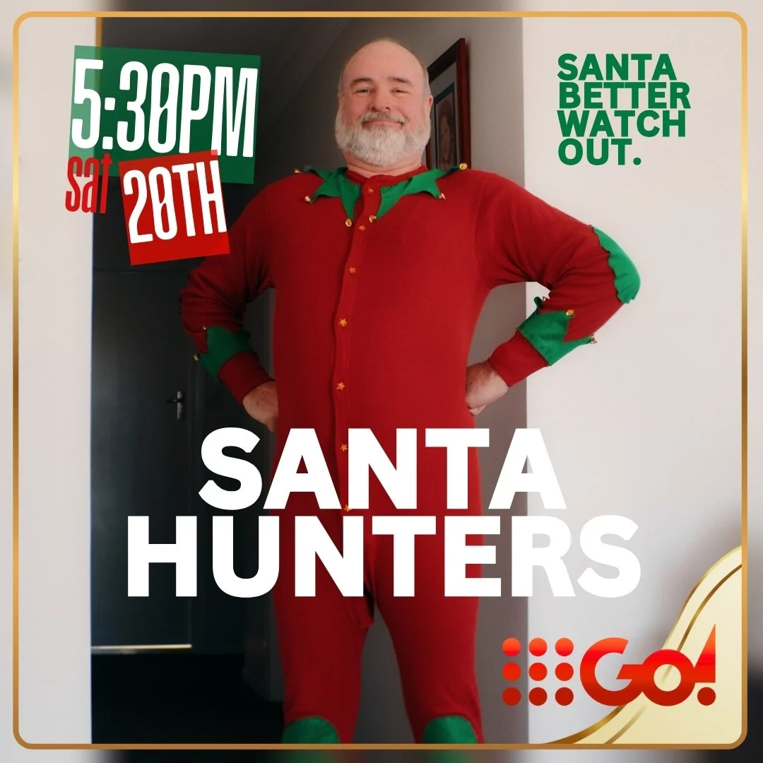 A man dressed in a red and green elf costume standing in a doorway, with one hand on his hip and smiling.