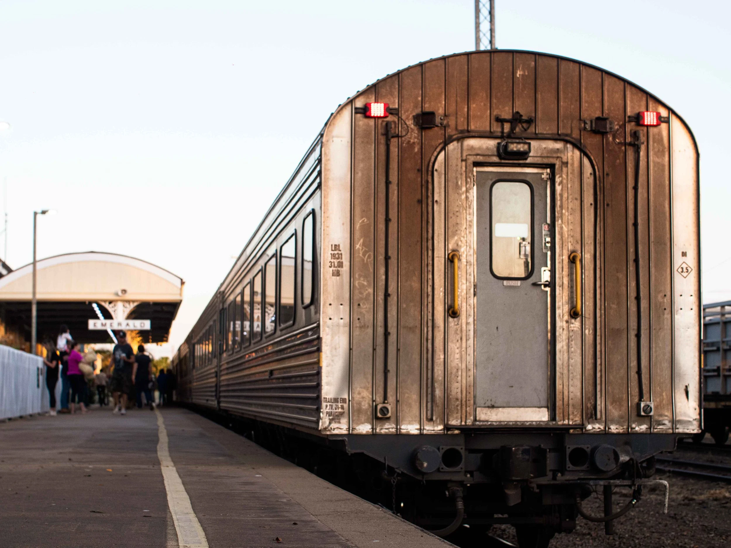 A vintage train car at a station platform with people waiting in the background, and a sign that reads 'EMERALD' under a covered area.