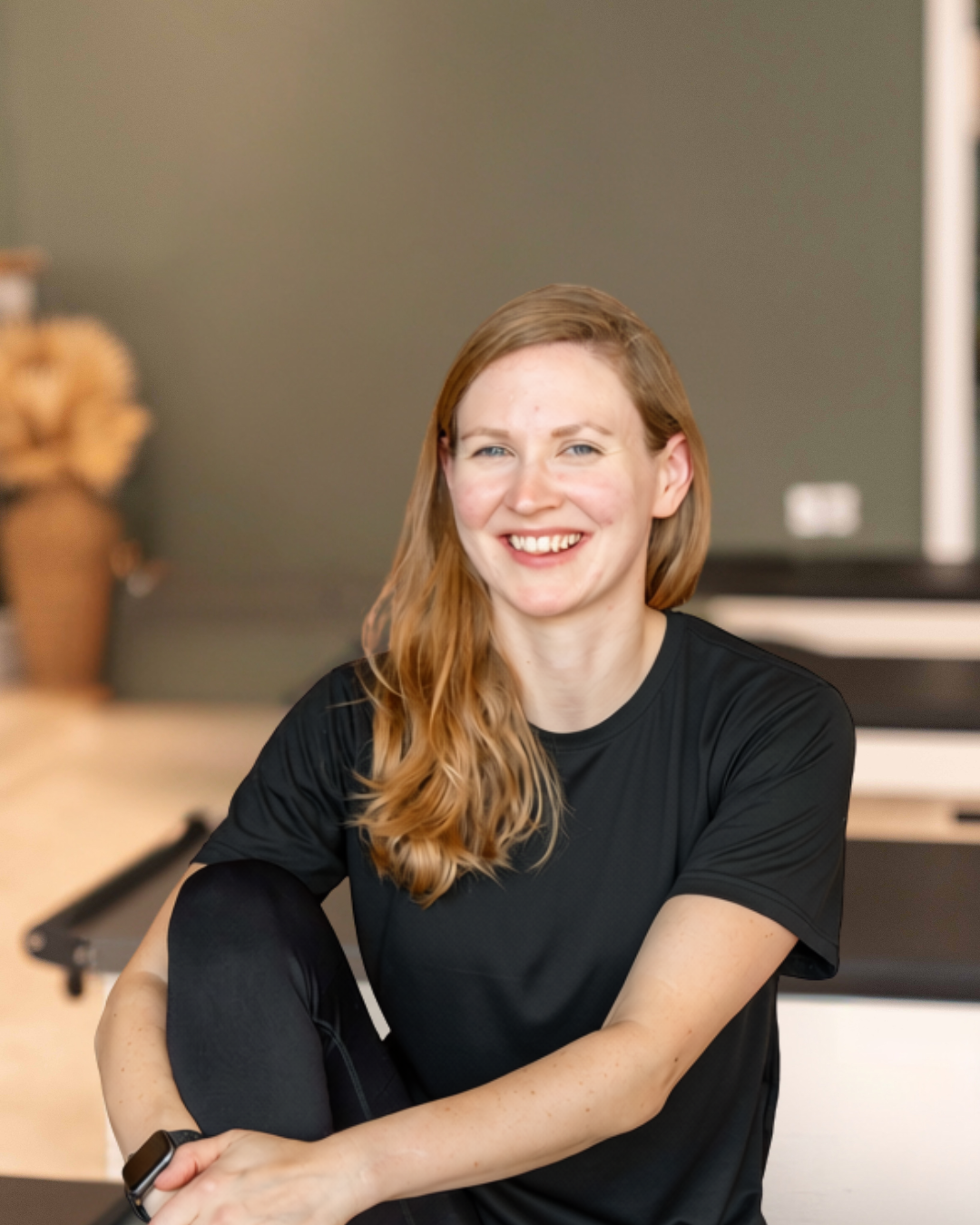 A woman with long red hair and a black athletic shirt sitting and smiling indoors.