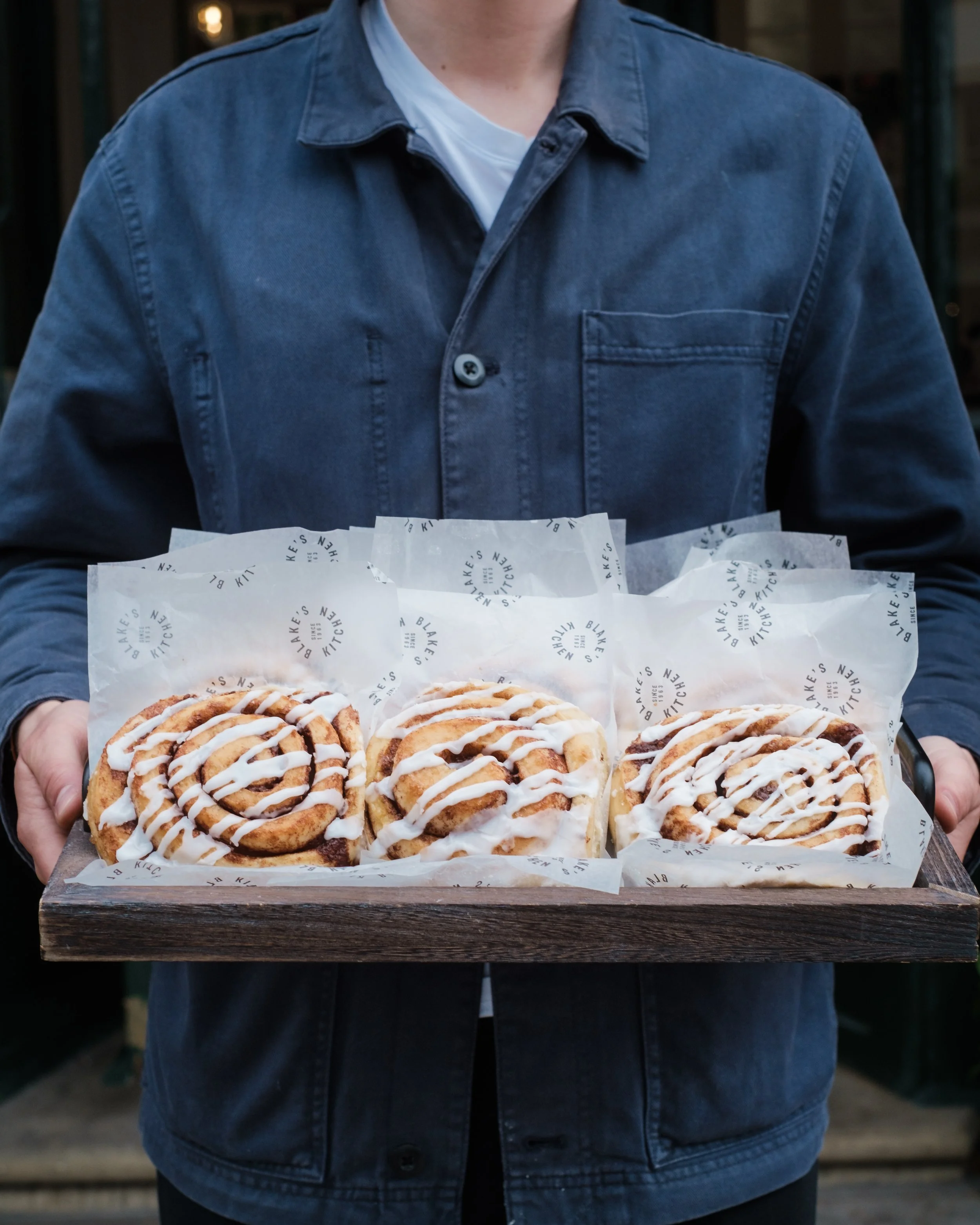 Man in blue shirt holding a tray of award-winning cinnamon buns from a family-run Cotswolds bakery, now served at Hagen in London.