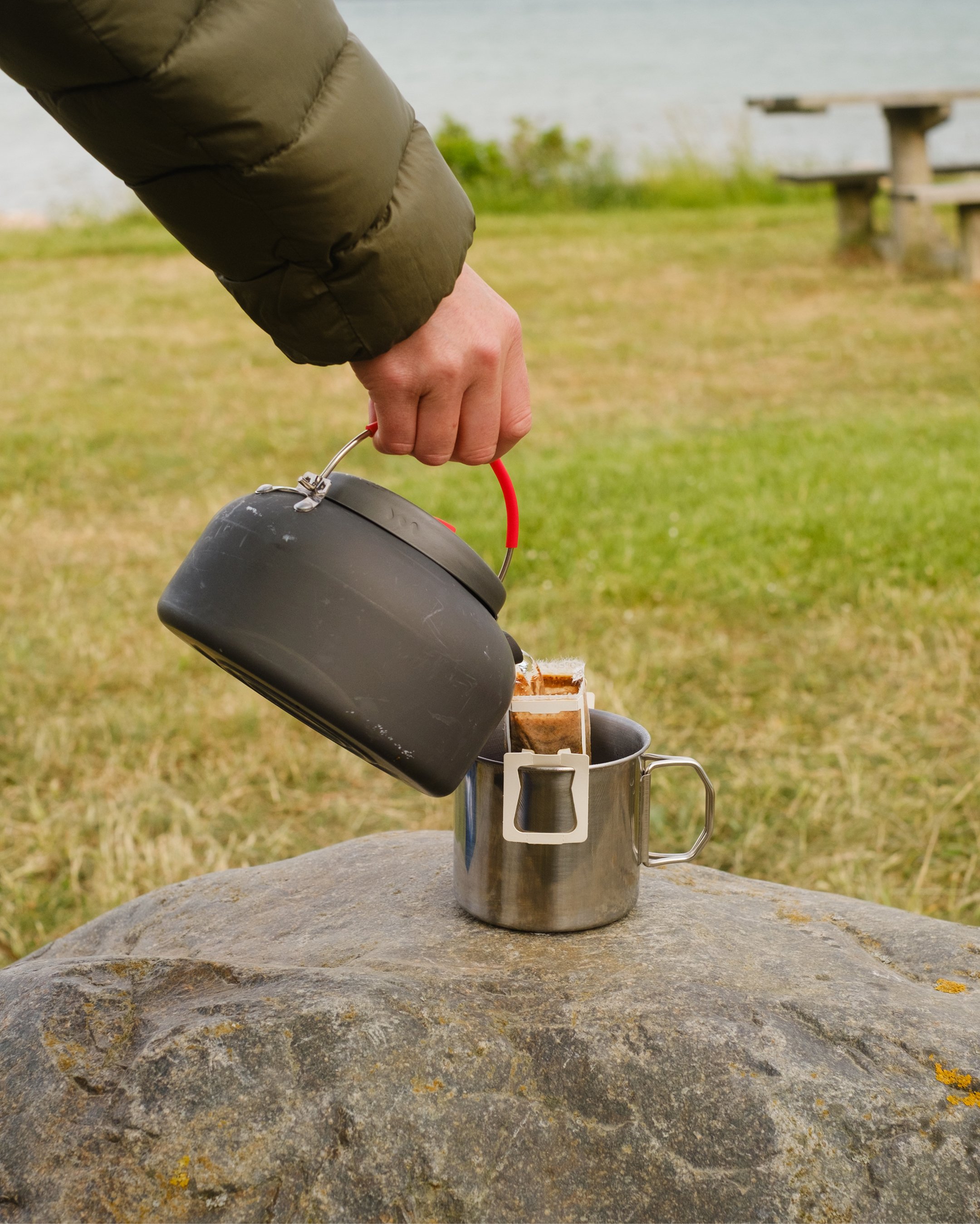 A person wearing a green jacket is pouring hot water into a metal mug with a coffee bag inside, outdoors on a large rock with grass and a picnic table in the background.