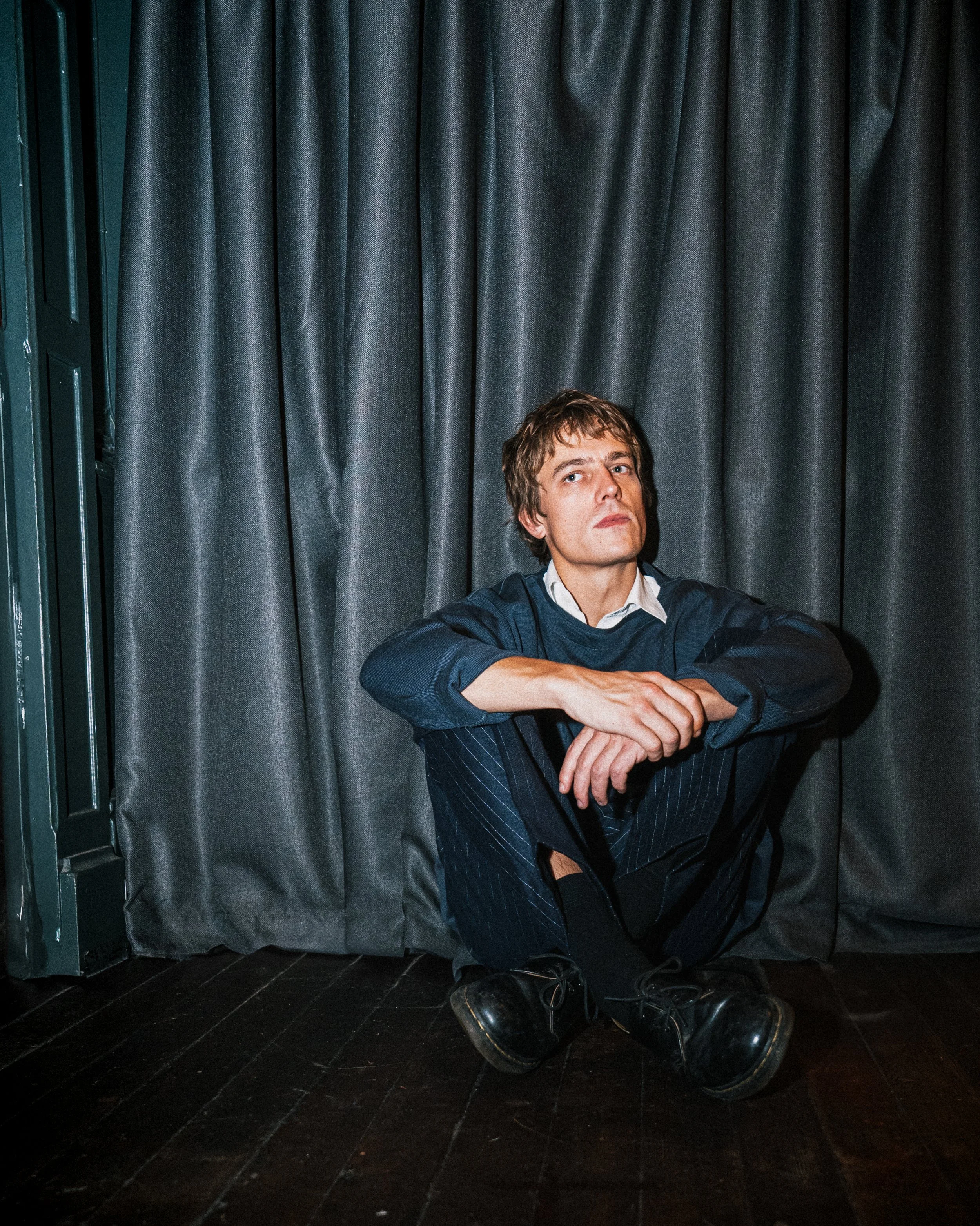 A man with brown hair sitting on a dark wooden floor against a gray curtain. He is wearing a navy blue sweater and pinstripe pants, with a somber expression.