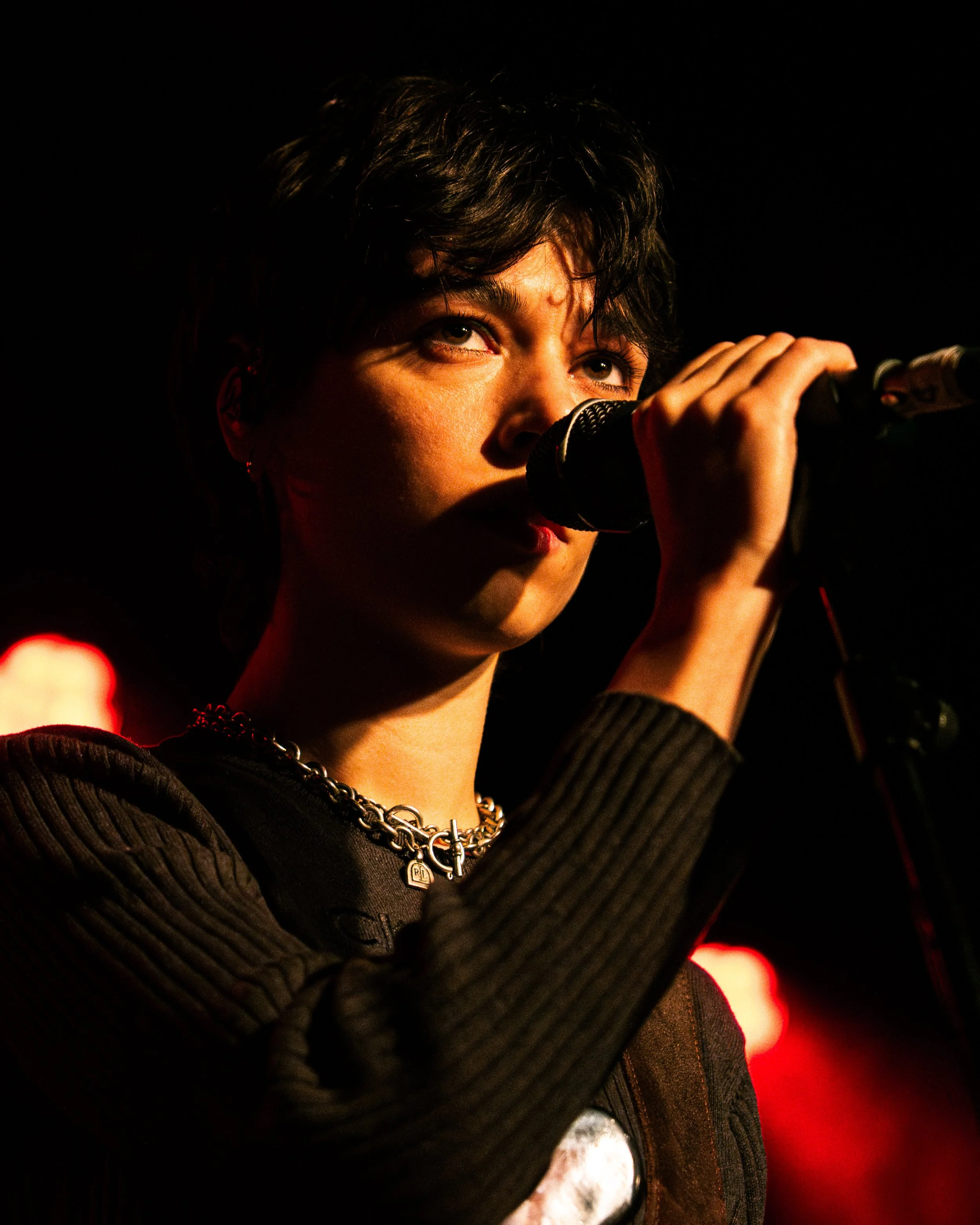 A person with short, curly dark hair singing into a microphone on stage, illuminated by warm stage lighting.