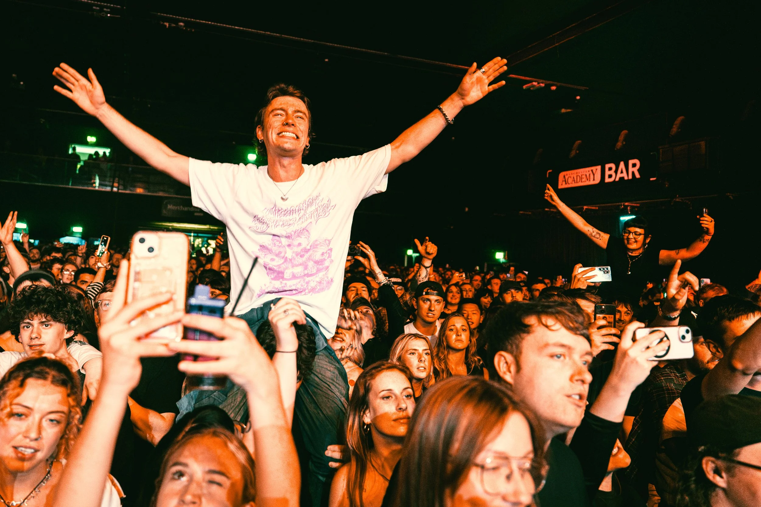 A person stands on someone's shoulders with arms outstretched at a concert, surrounded by an audience taking photos and videos.