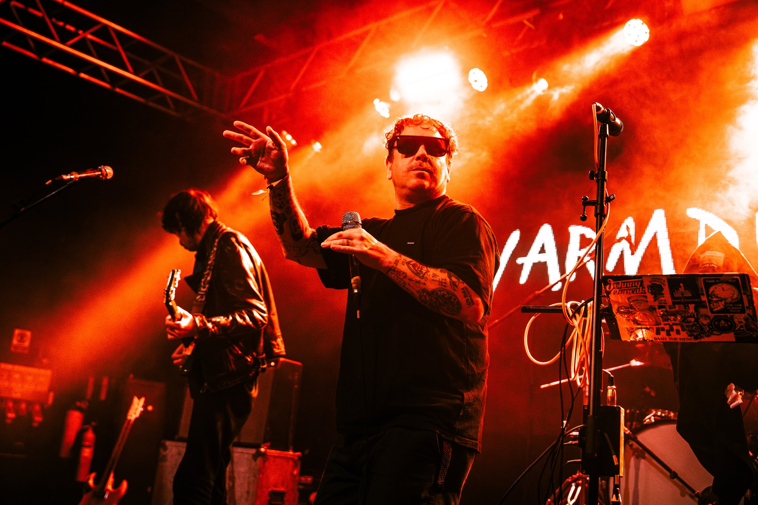 A band performs on stage with orange lighting. The singer, with tattoos and sunglasses, holds a microphone and gestures with his hand. Another musician plays guitar in the background.