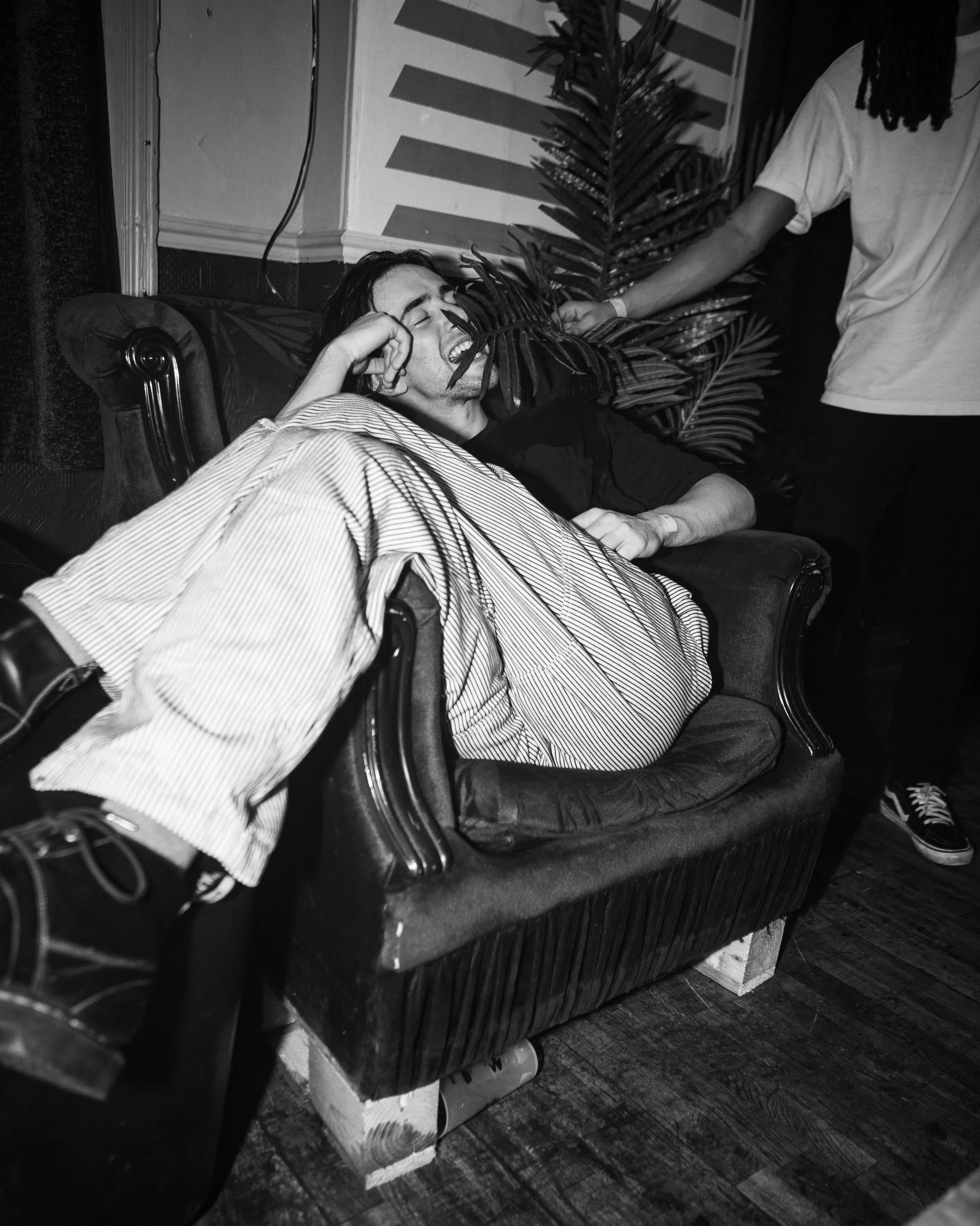 A man with dreadlocks and striped pants is reclining on a vintage armchair, laughing with his eyes closed. Another person in a white T-shirt is partially visible, touching a large plant behind him. The setting appears to be indoors with wooden floori