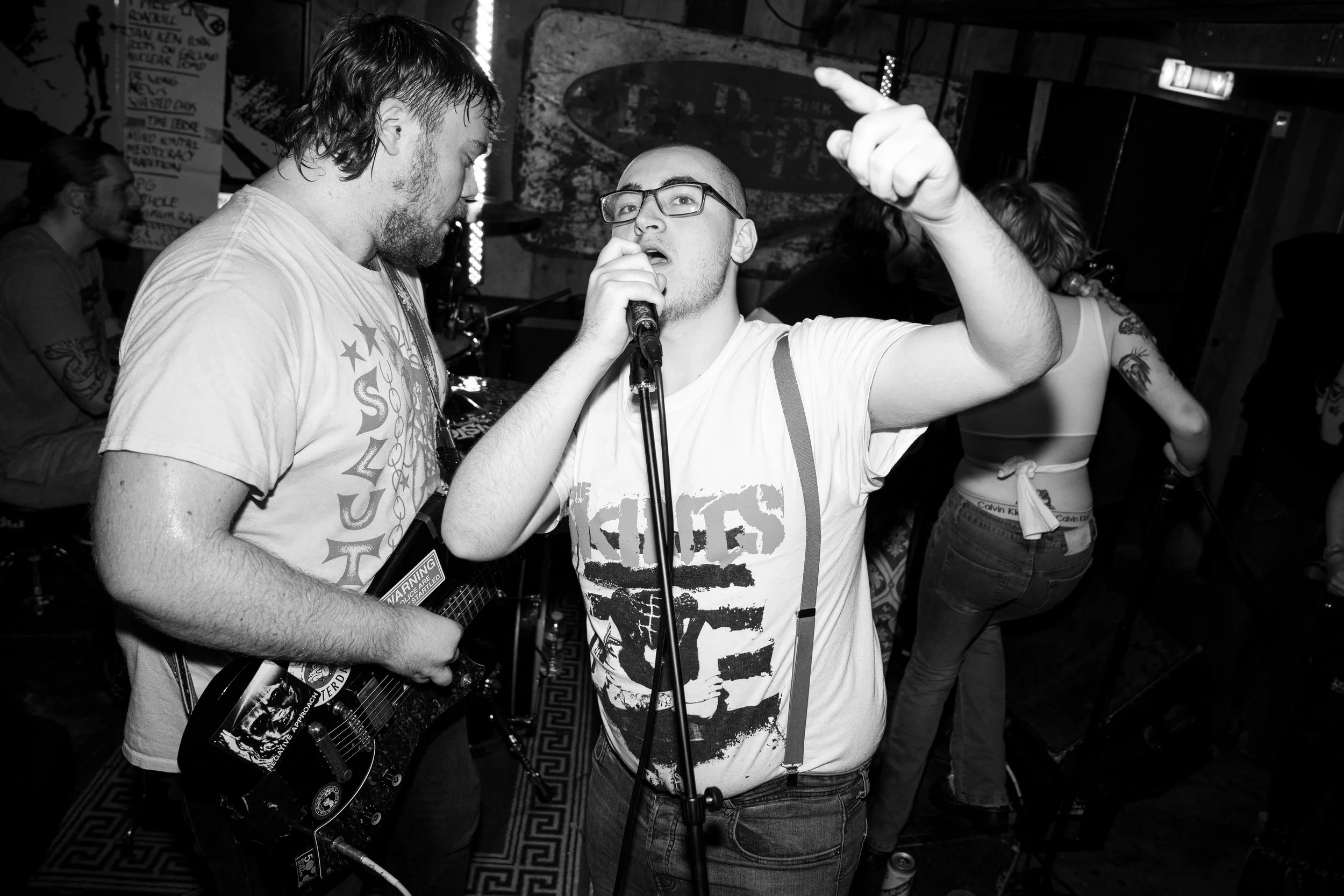 A black and white photo of a live music performance in a small venue. A man with glasses and a graphic T-shirt sings into a microphone with one hand raised, while another man plays an electric guitar next to him. In the background, there are two wome
