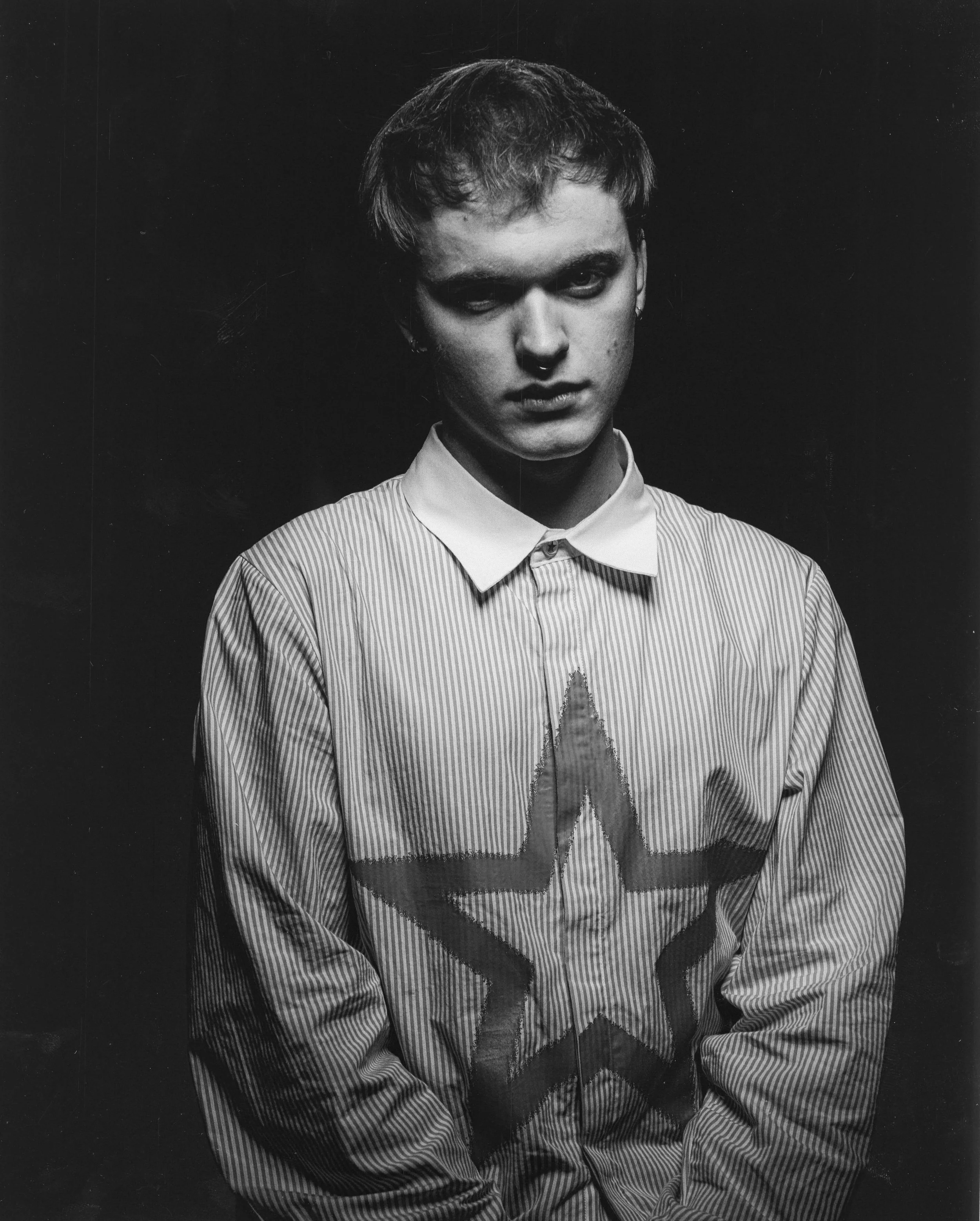 Black and white portrait of a young man with tousled hair, wearing a striped shirt with a large star on it, against a dark background.