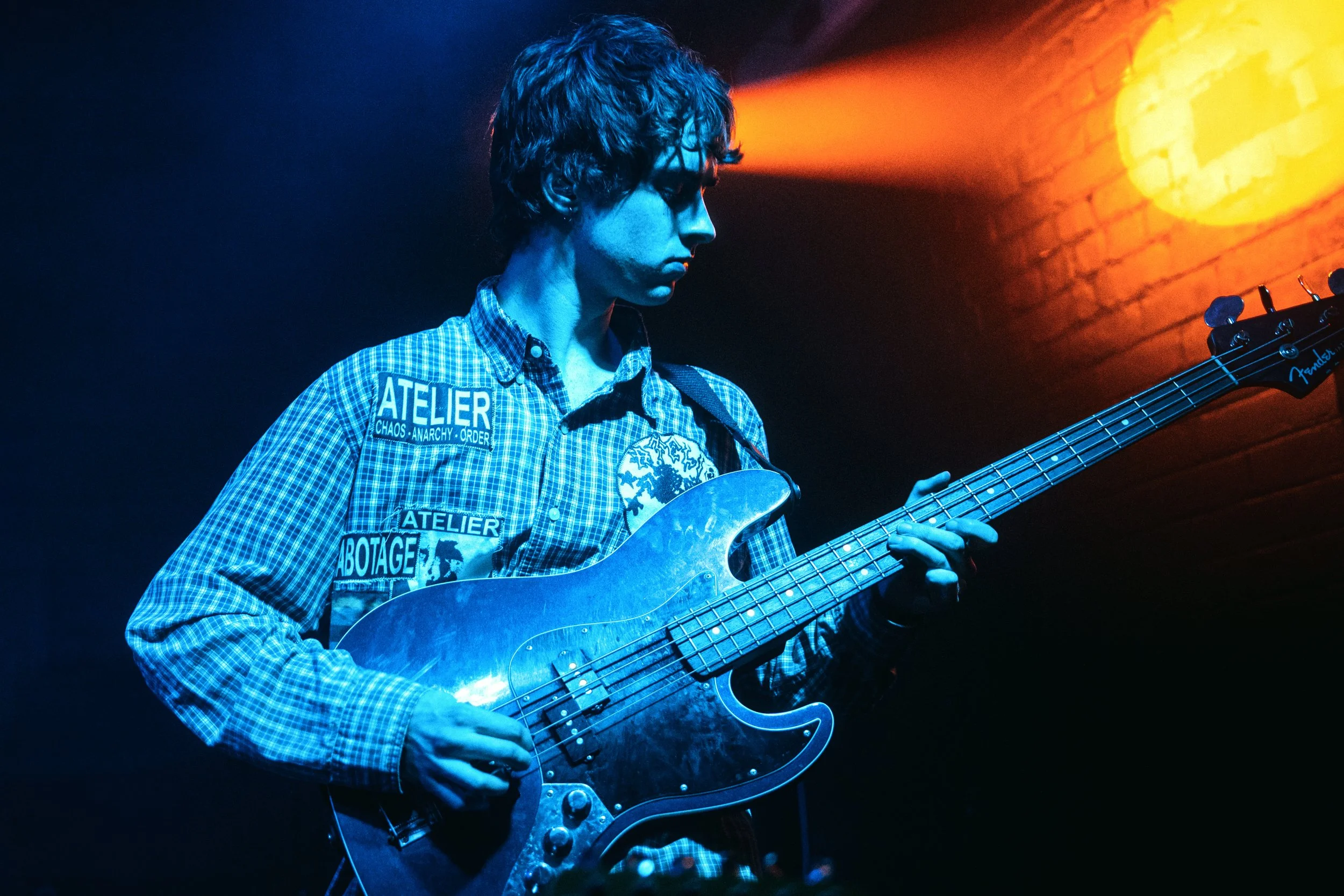 A person with curly hair playing a Fender electric bass guitar on stage, wearing a checkered shirt with patches, under blue and orange stage lighting.