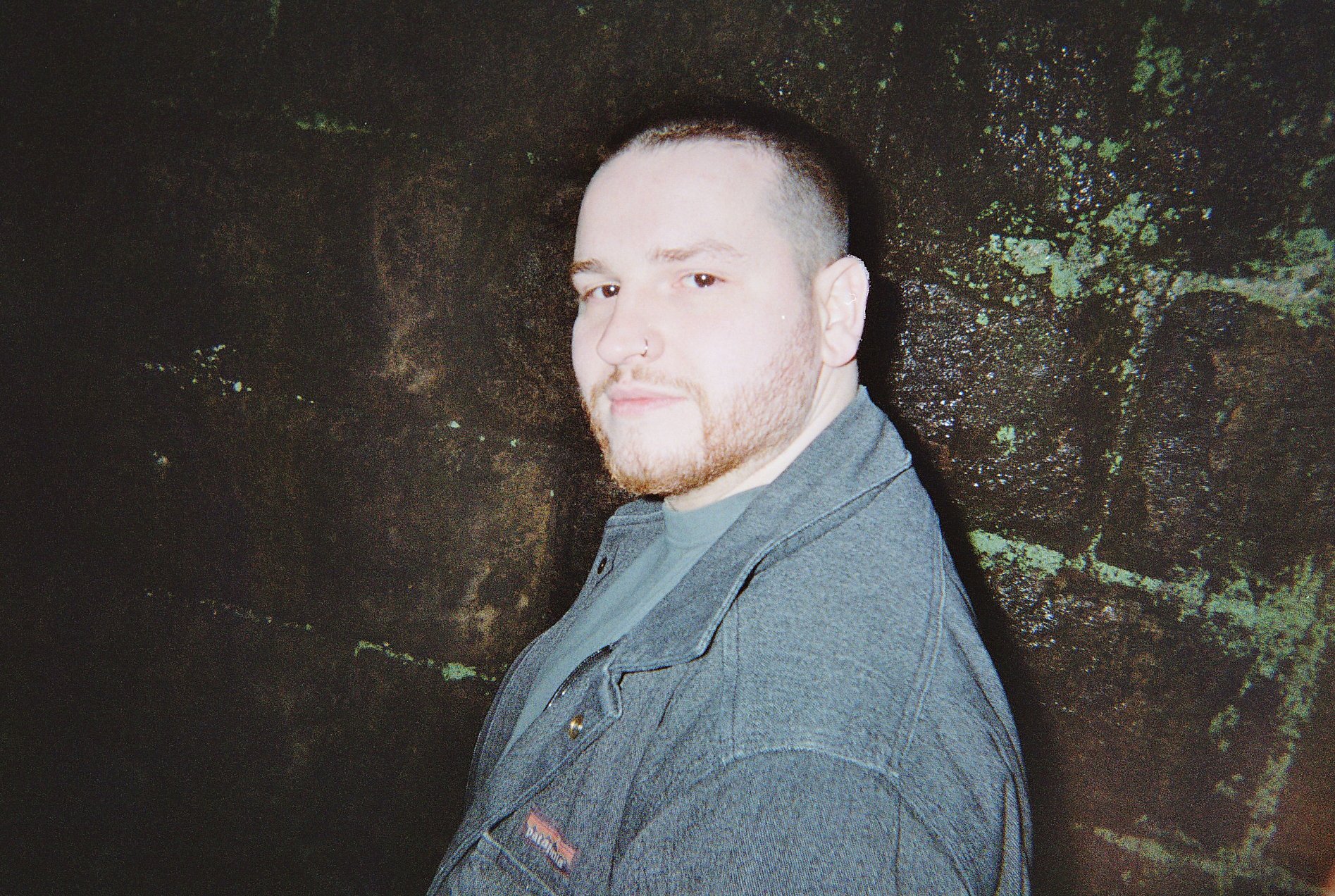 A man with a beard and mustache, wearing a gray jacket, stands against a dark, textured wall.