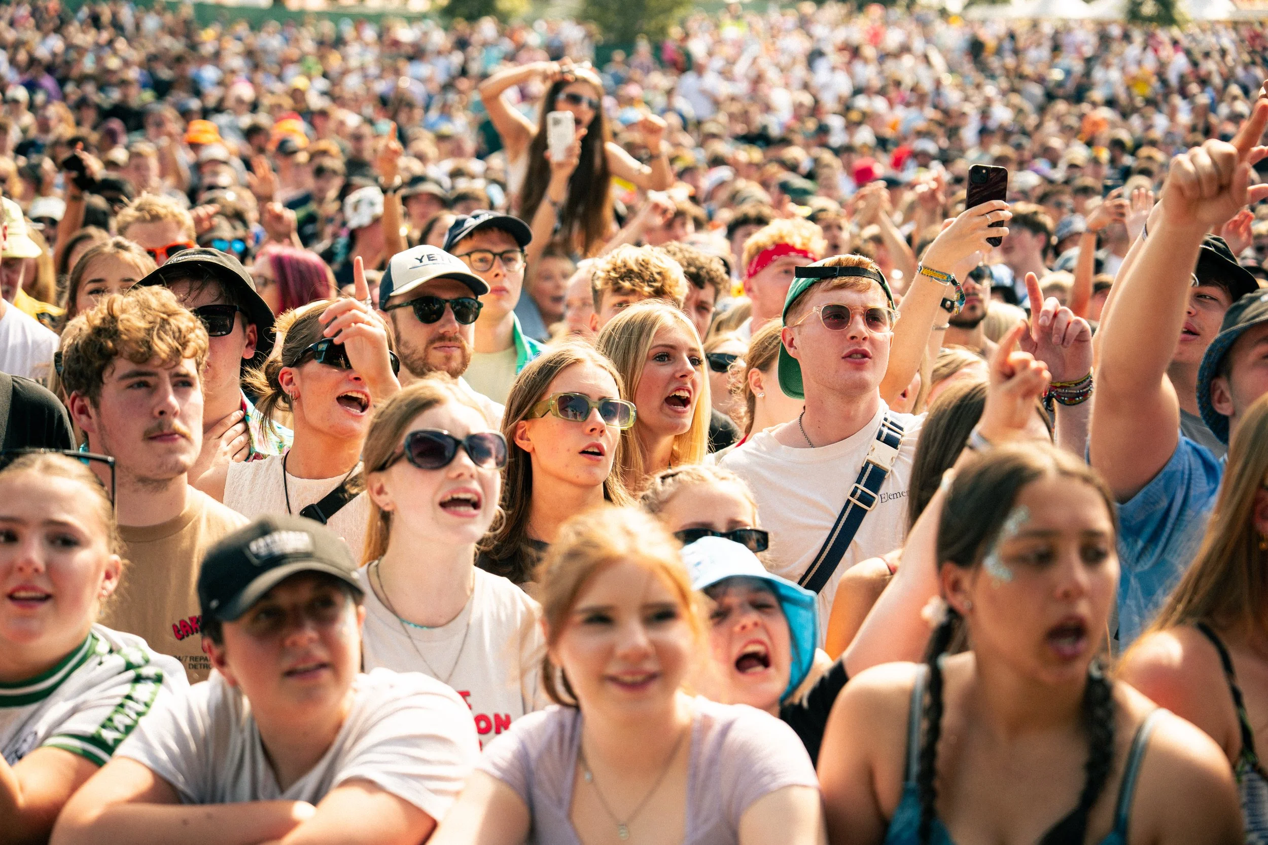 Crowd of young people at an outdoor concert or festival, some cheering, singing, and using their phones to record or take pictures.
