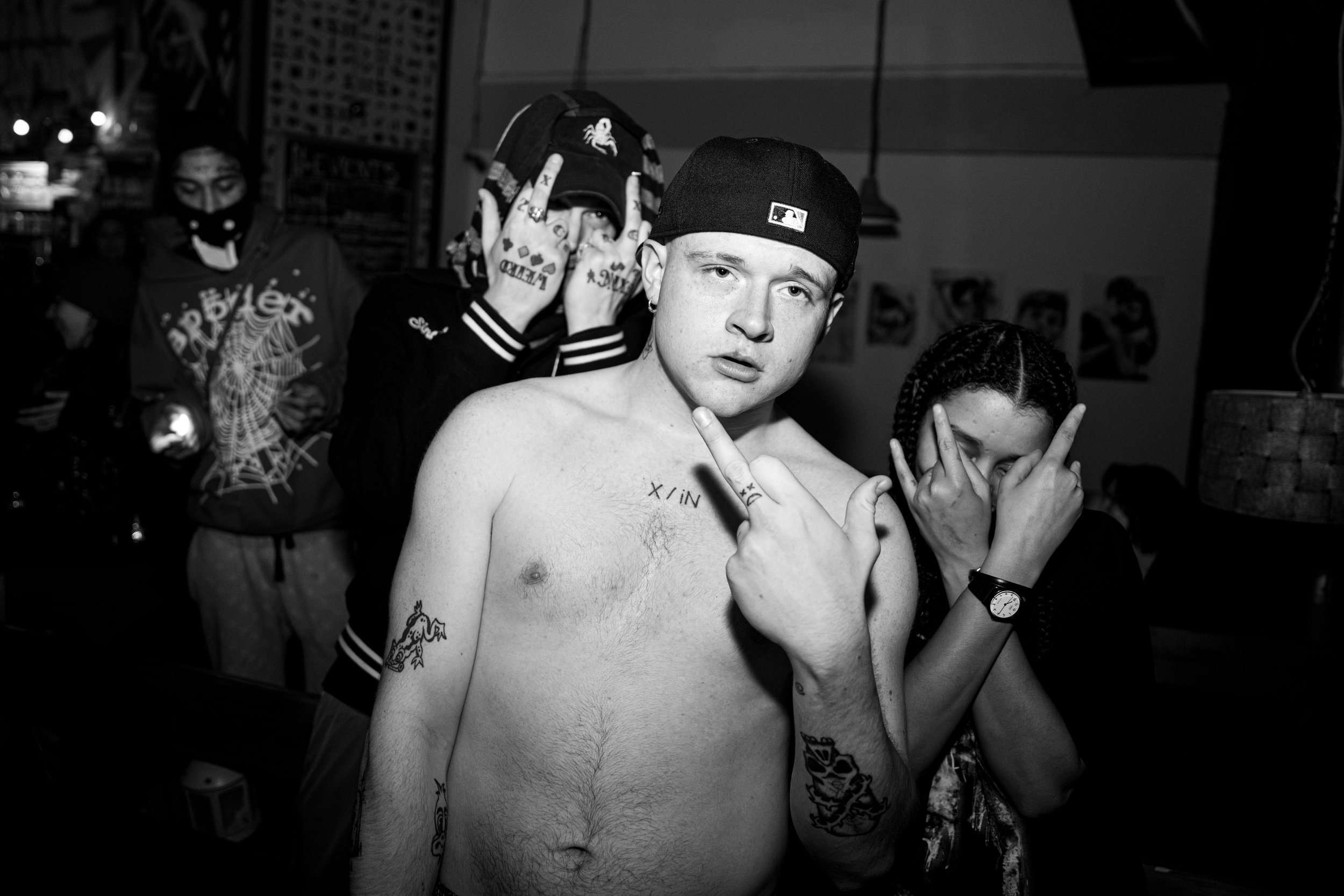 Black and white photo of four young people at a party or club. The shirtless young man in the foreground wears a cap backward, has tattoos, and makes a hand gesture. The young woman next to him covers her face with her hands. Two individuals are in t
