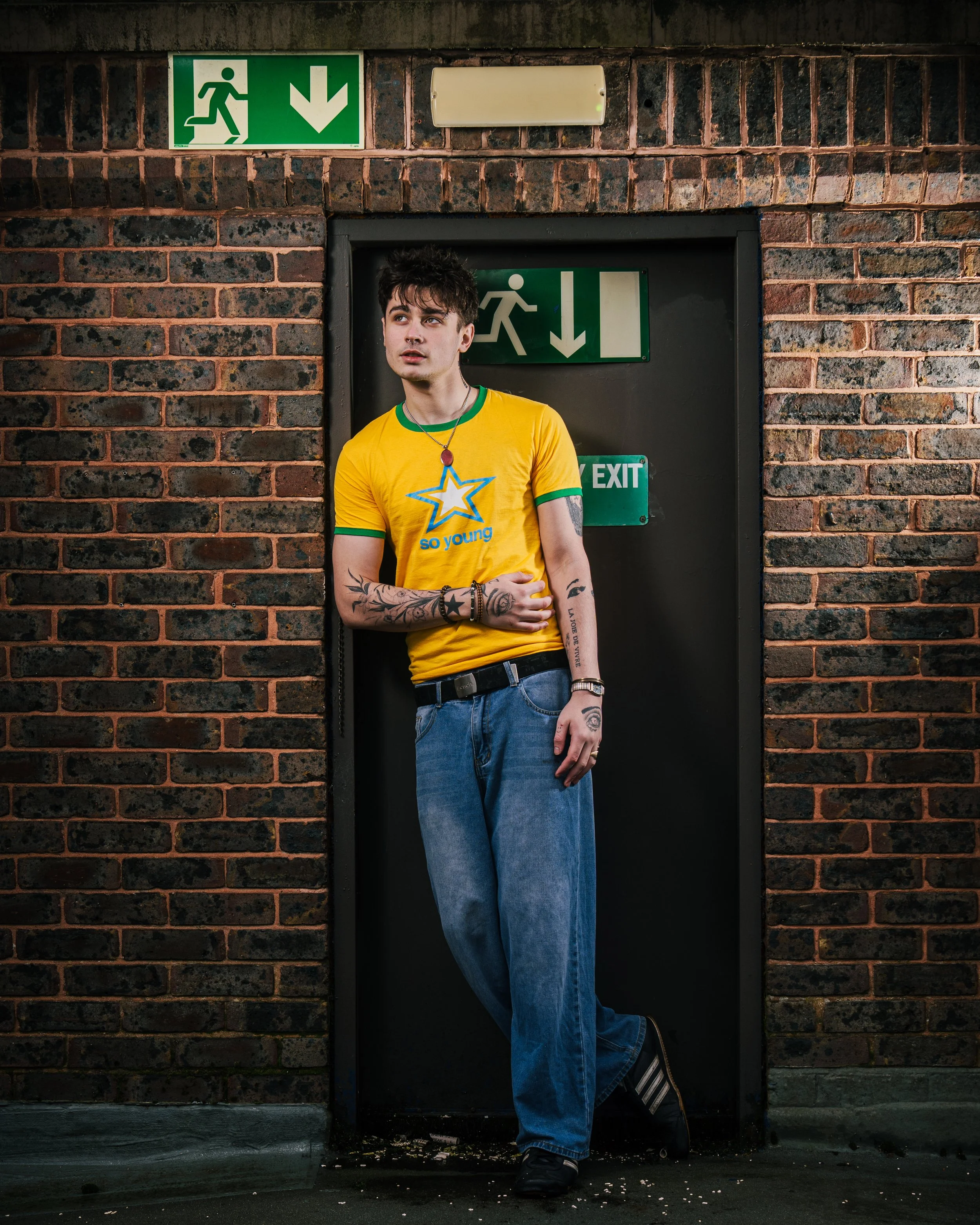 Young man with tattoos and casual clothing standing in front of a black emergency exit door with green and white exit signs, against a brick wall.