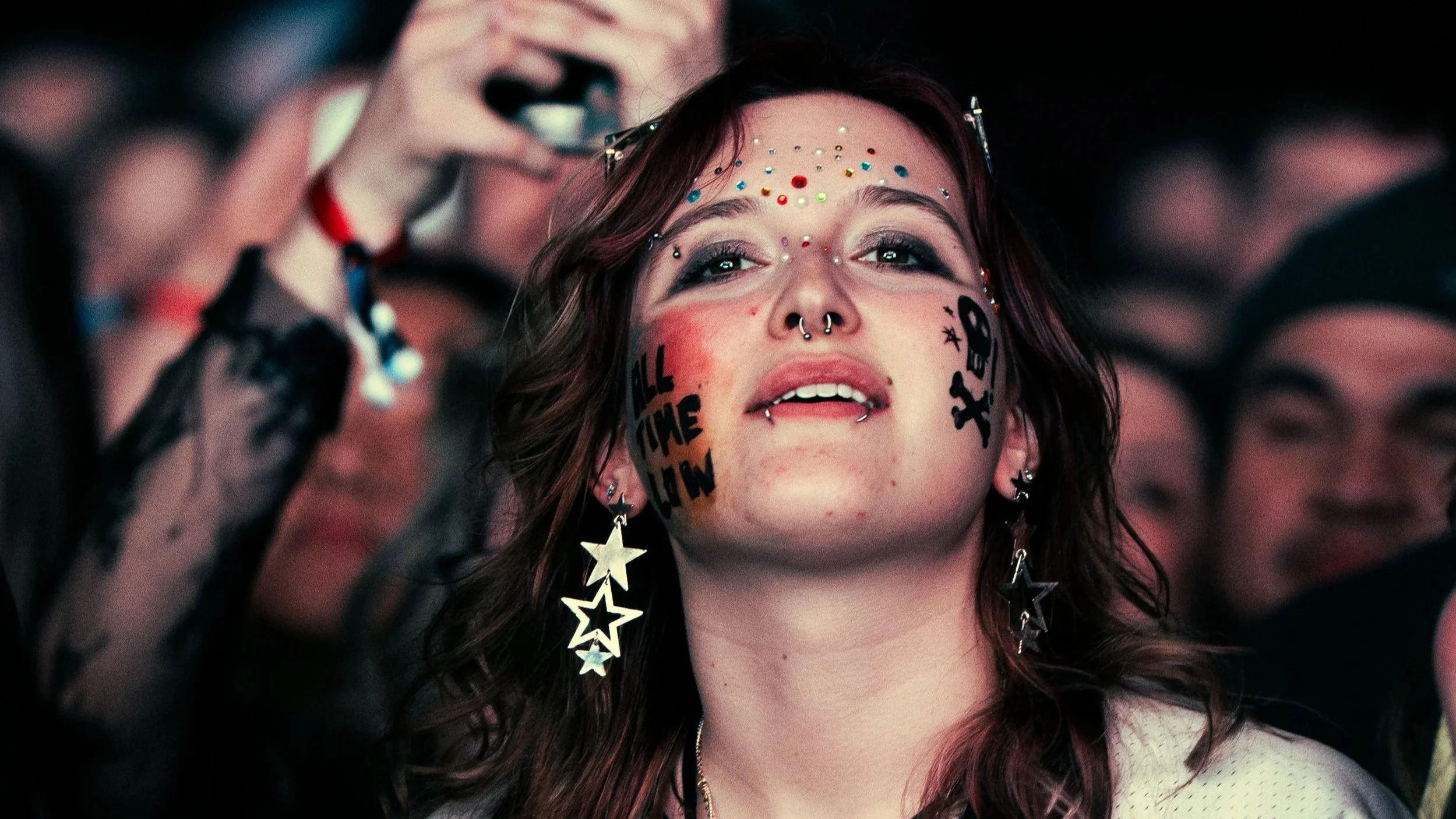 A young woman with colorful facial stickers and face tattoos smiling at a concert or festival. She has star-shaped earrings, septum and lip piercings, and dark hair with purple highlights. She is surrounded by a crowd, some of whom are taking picture