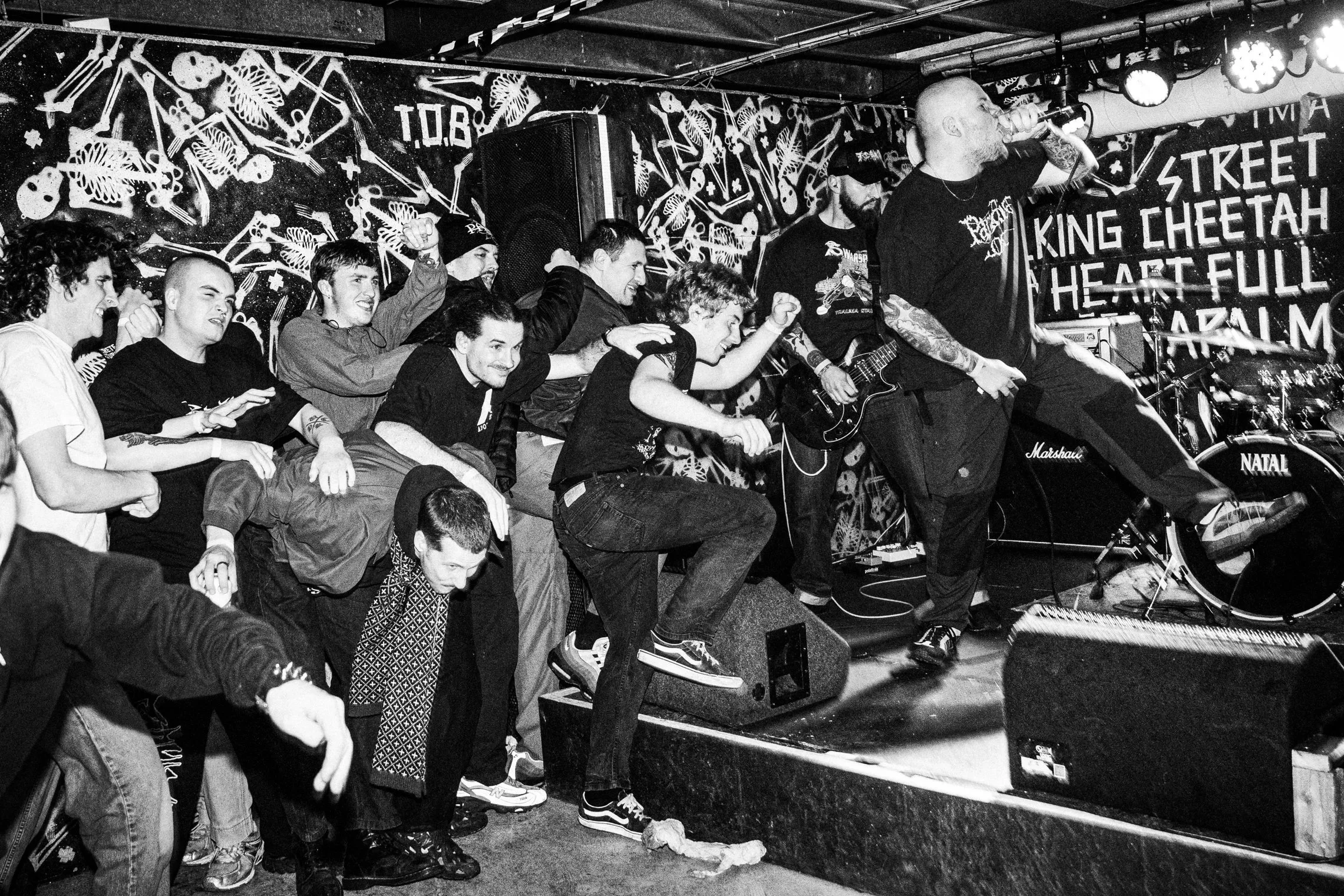 A black and white photo of a male singer performing on stage with a band, engaging with a crowd that is moshing and jumping, at a small music venue with graffiti art on the wall behind them.