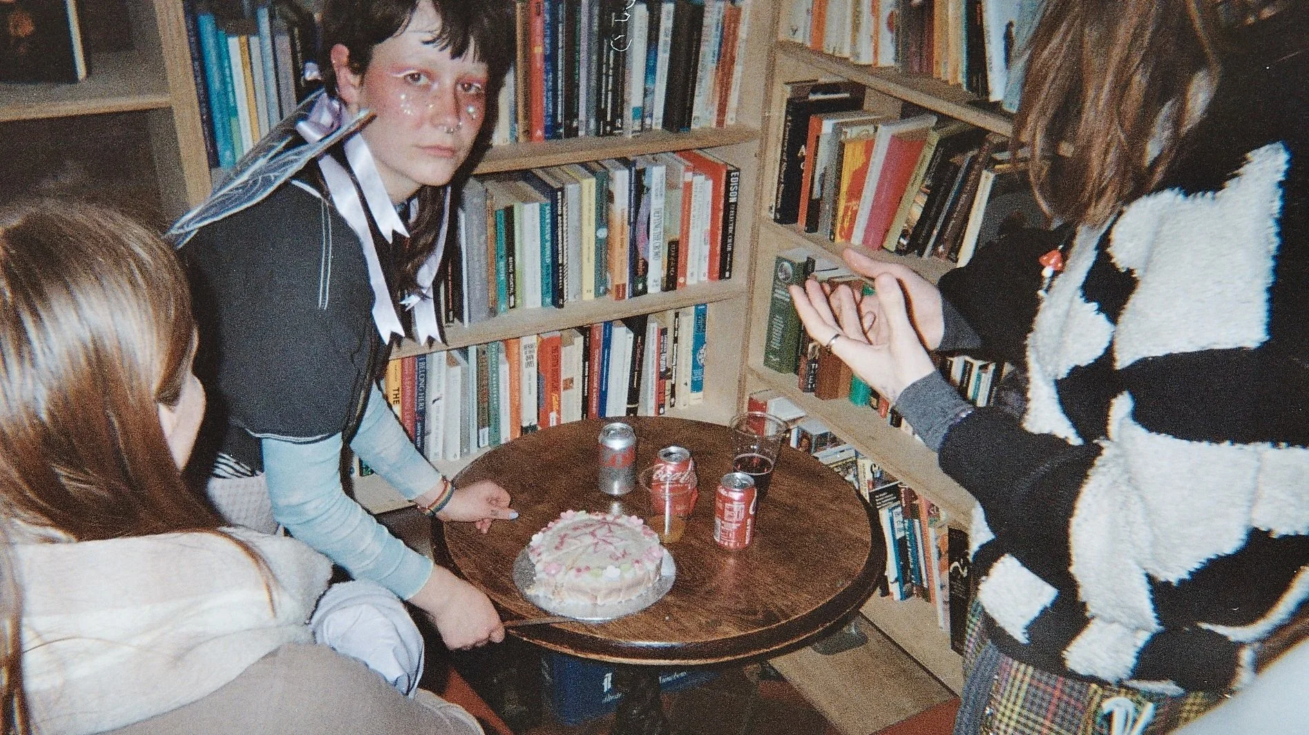 Three young women gathered around a small wooden table with a decorated birthday cake and soda cans, in front of a bookshelf filled with books. One woman is looking directly at the camera, while the other two are partially visible.
