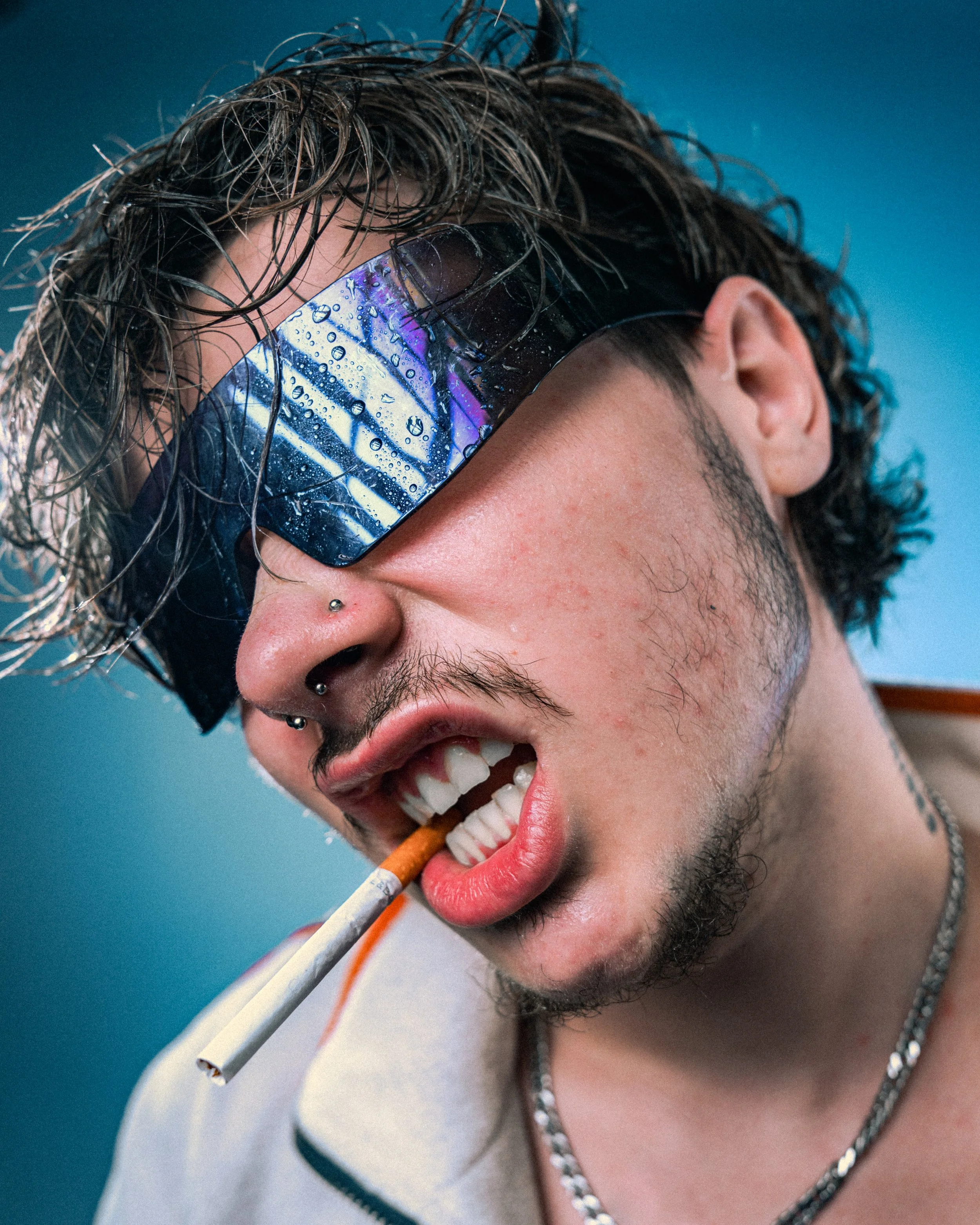 Close-up of a young man with wet hair wearing reflective sunglasses, biting a cigarette, and showing a rebellious attitude.