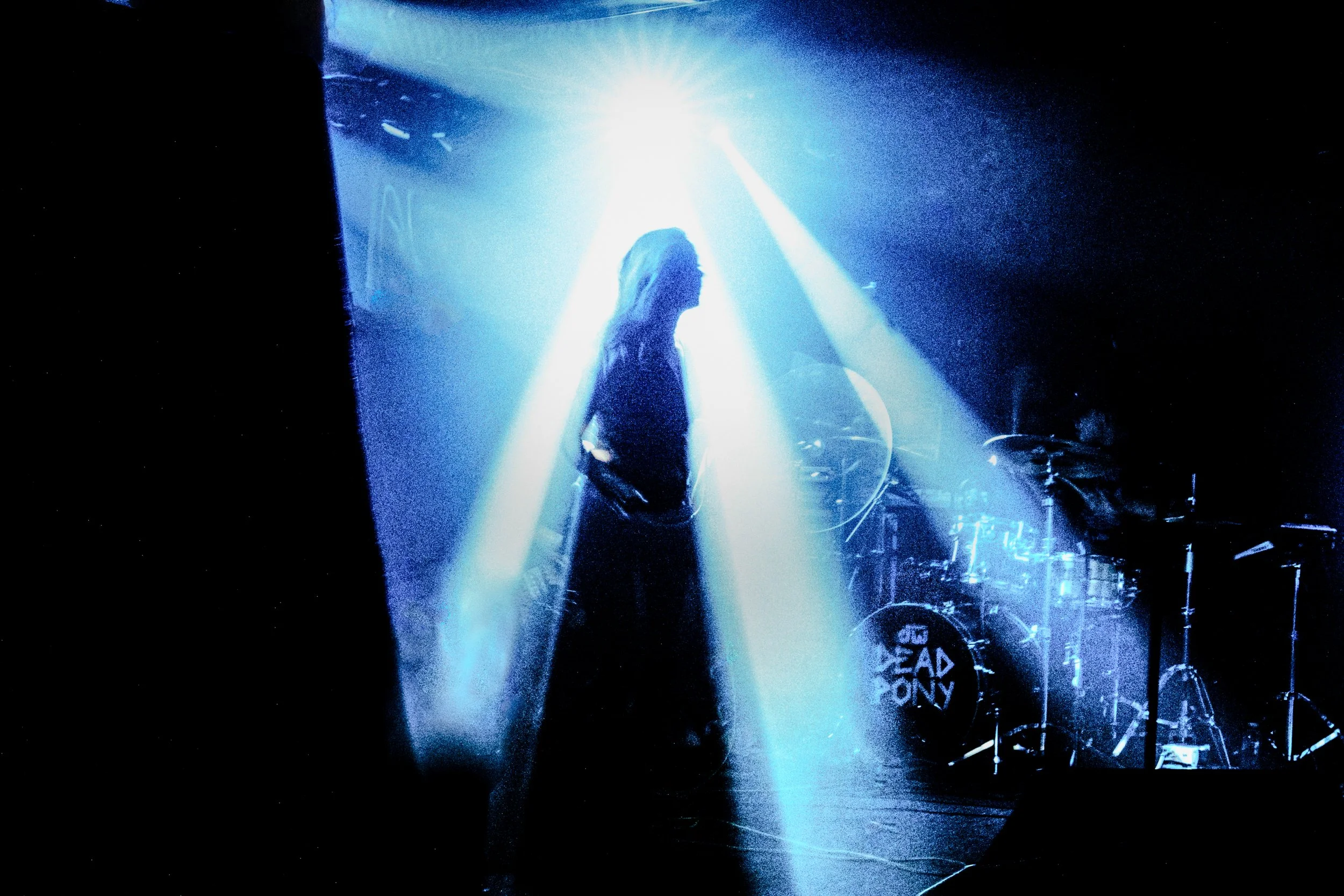 Silhouette of a woman standing in a dark stage, illuminated by bright white and blue spotlight beams, with a drum set in the background.