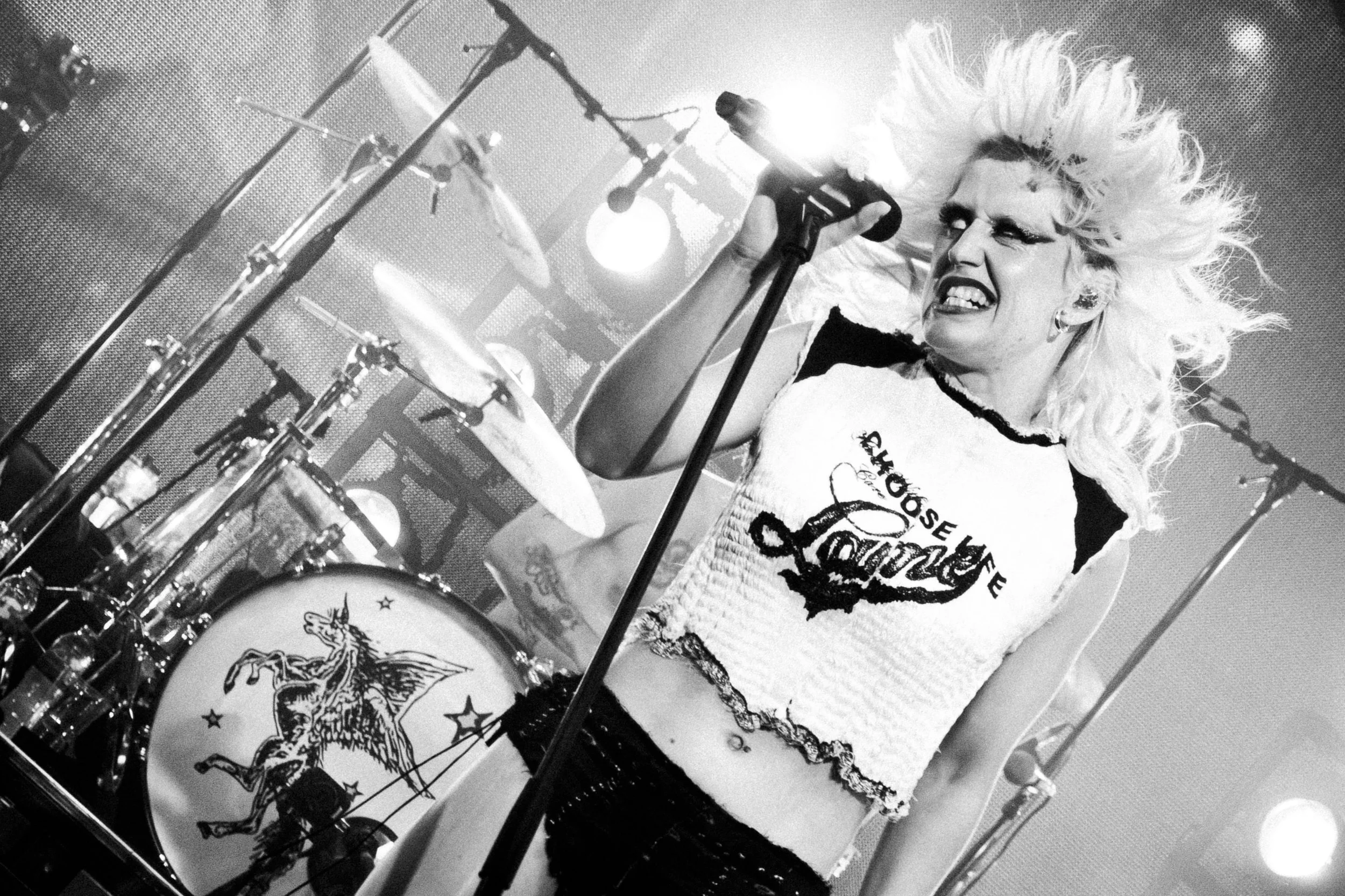A woman performing on stage with a drum set, holding a microphone in her right hand, with a fierce expression, and wearing a sleeveless top with text and graphics.