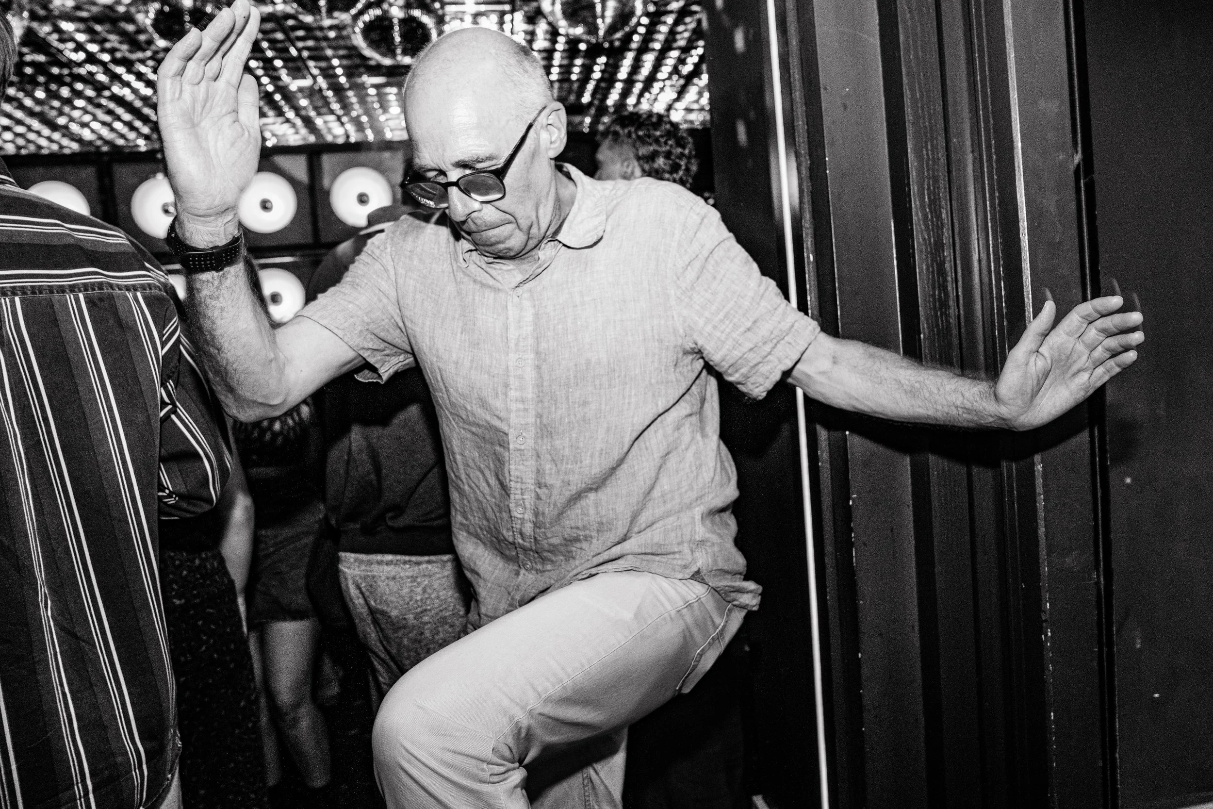 An elderly man with glasses dancing with one foot raised, eyes closed, and arms outstretched in a nightclub or dance hall with a patterned ceiling and other people in the background.