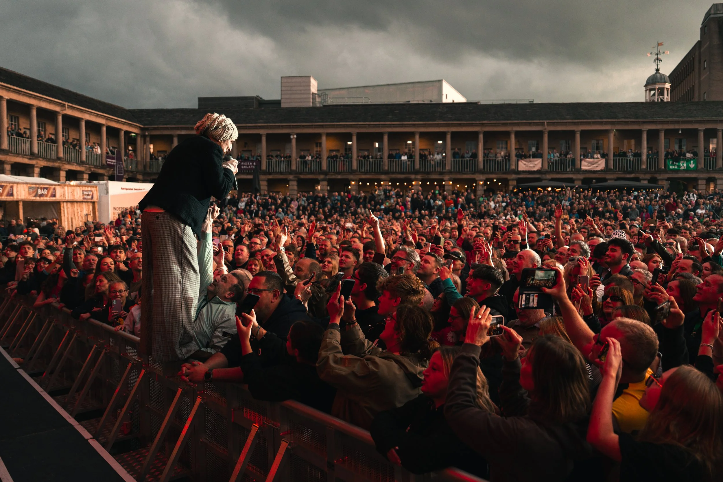 Large outdoor concert with dense crowd of people, performer on stage, and audience taking photos.