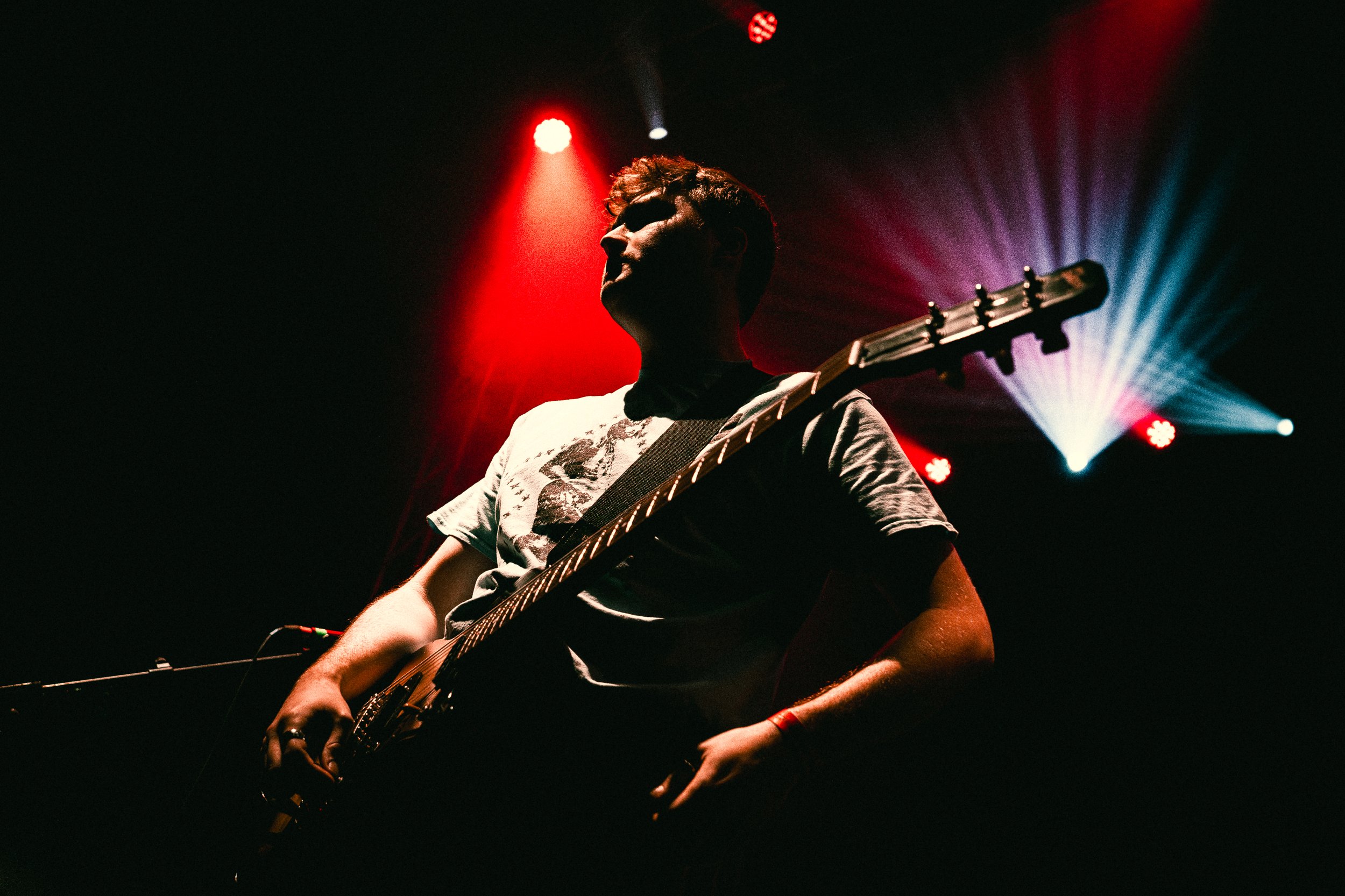 A person playing an electric guitar on stage, illuminated by red and white stage lights, with a dark background.