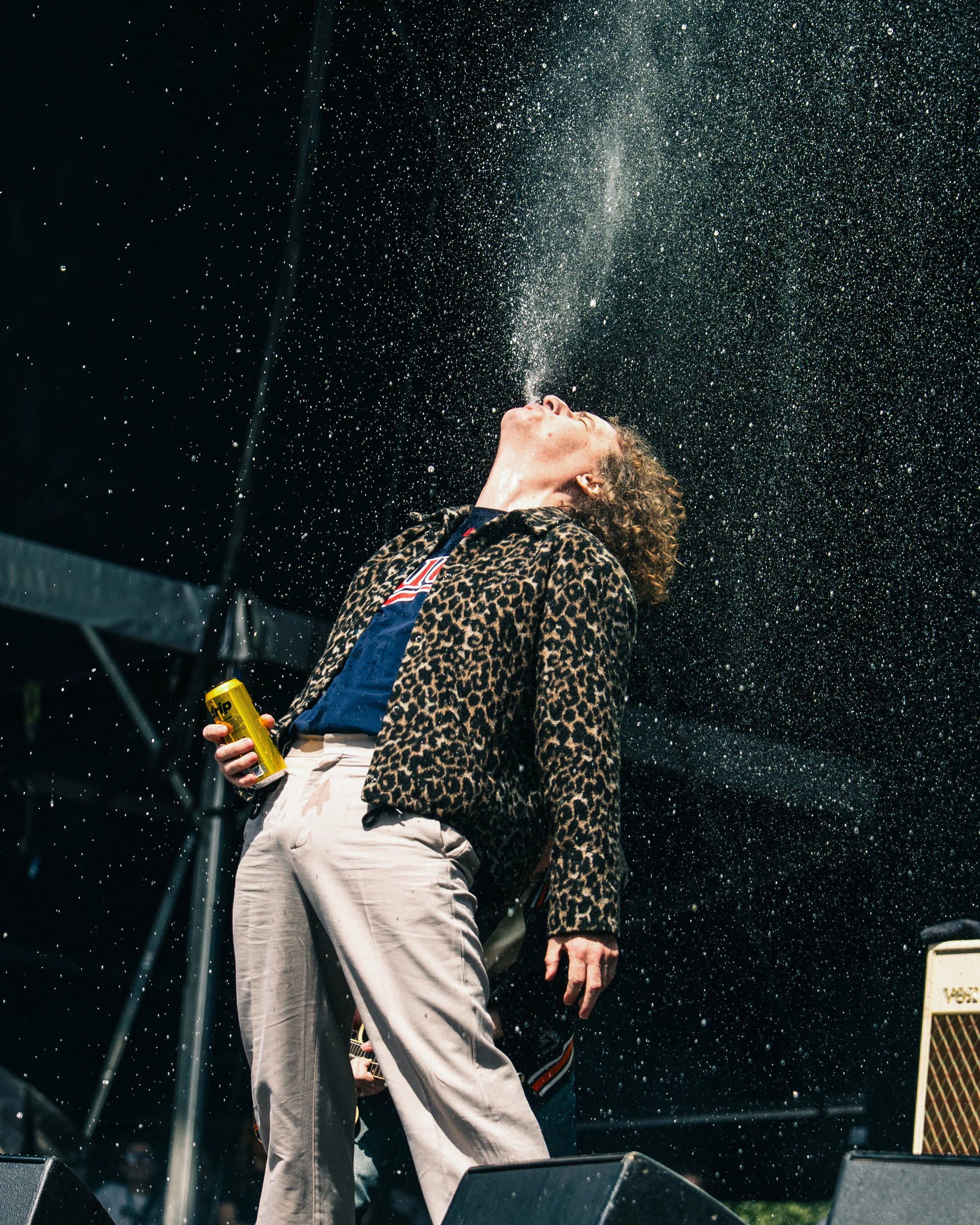 A person in a leopard print jacket and light-colored pants is standing on a stage, tilting their head back as water splashes from their mouth into the air. The person is holding a yellow can in their right hand, and there is a black background with s