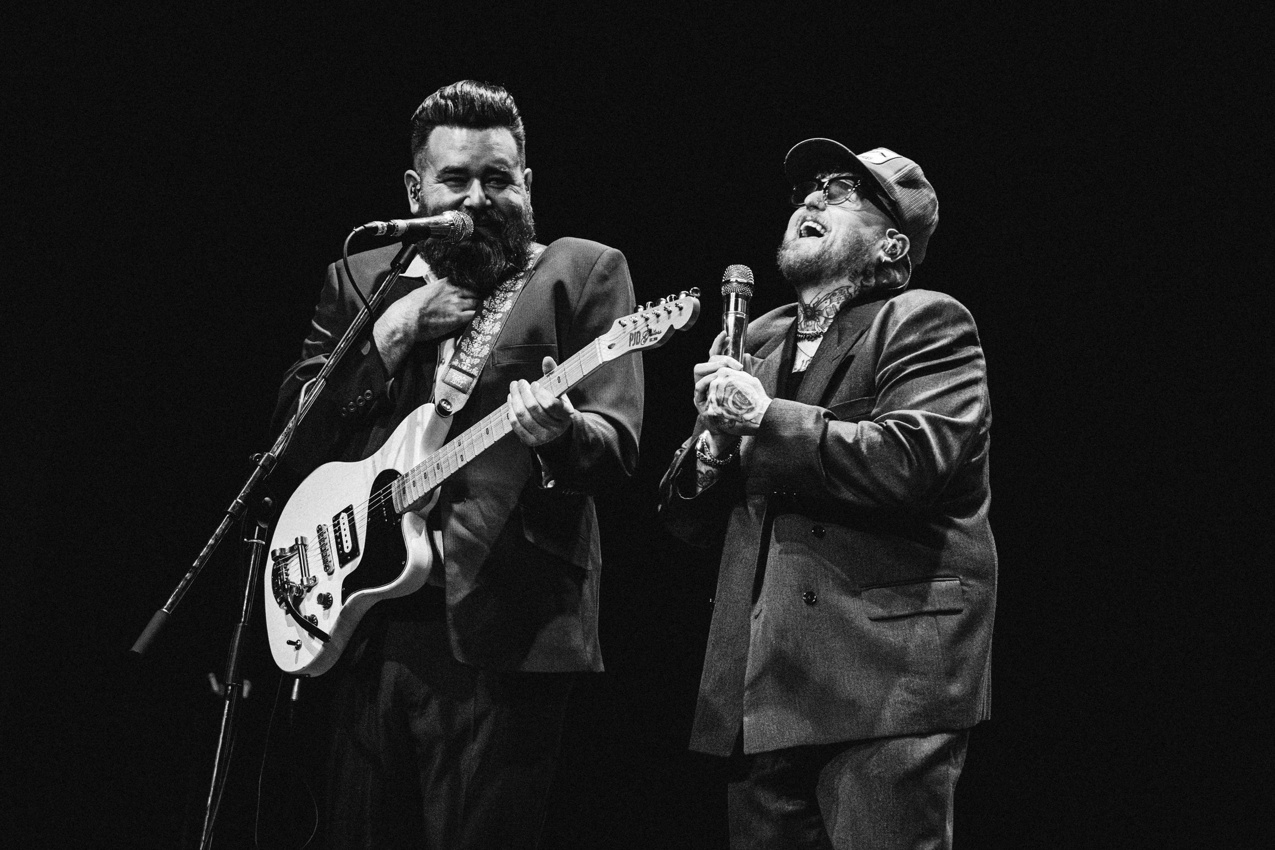Two men performing on stage, one playing an electric guitar and singing into a microphone, the other singing into a microphone with a cap and tattoos, both dressed in suits.