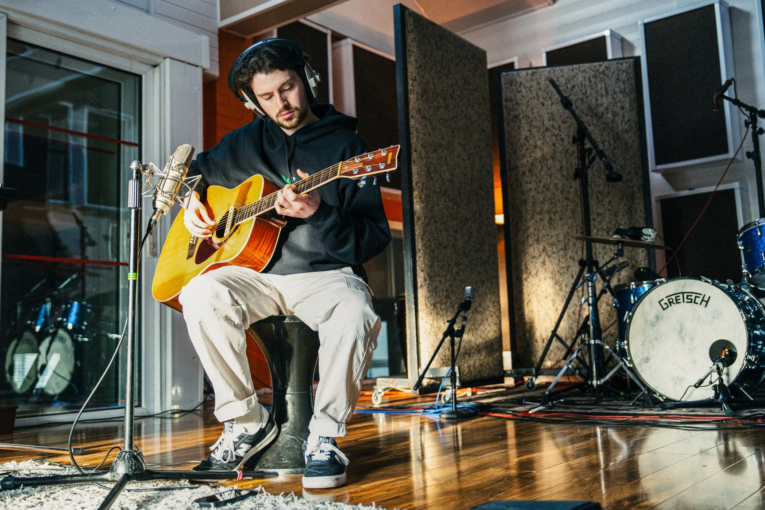 A young man with dark hair and a beard, wearing headphones, a black hoodie, and beige pants, sits on a black stool playing an acoustic guitar in a recording studio. There is a professional microphone set up in front of him and a drum kit with a Grets