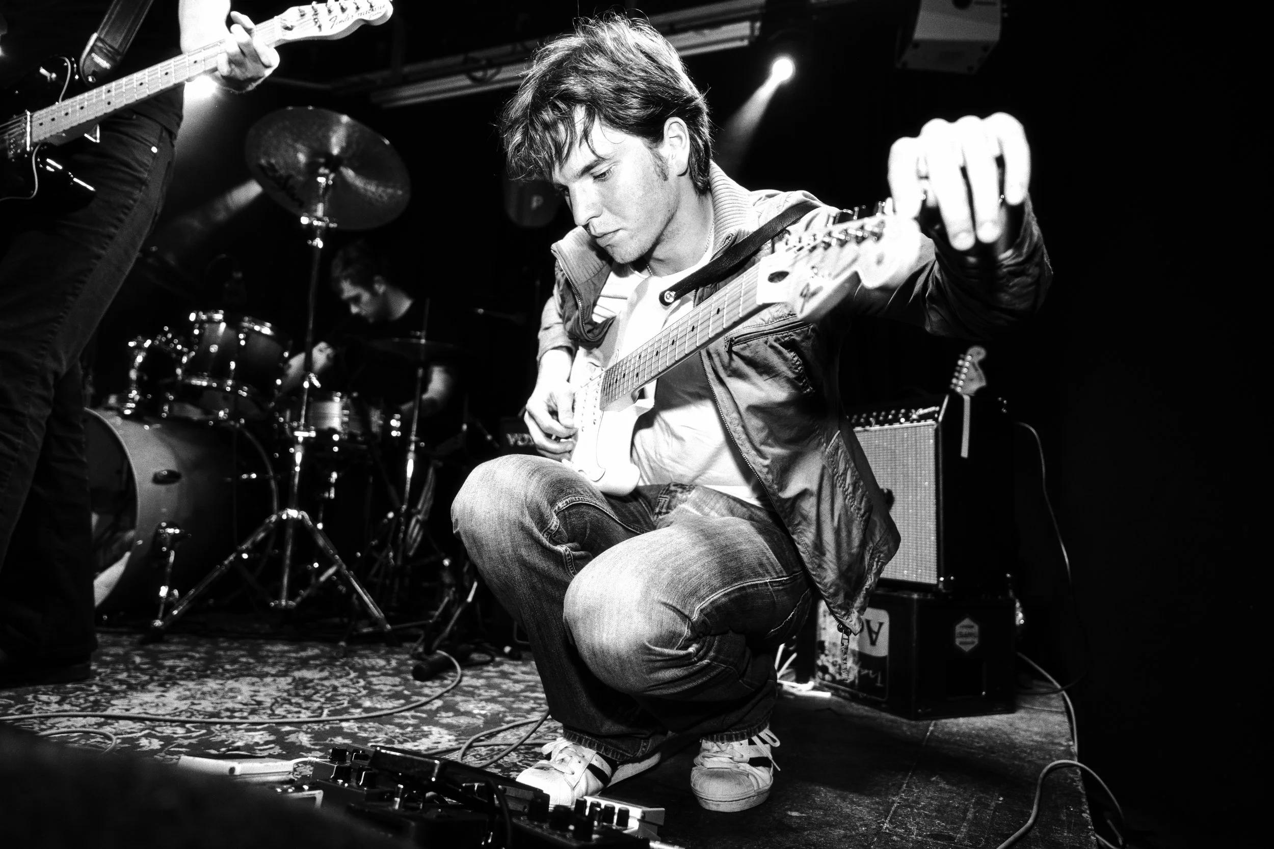 A black-and-white photo of a band performing live on stage. A young man is kneeling and playing an electric guitar, focusing on his instrument. Behind him, a drummer is playing drums, and another guitarist can be seen partially on the left side of th