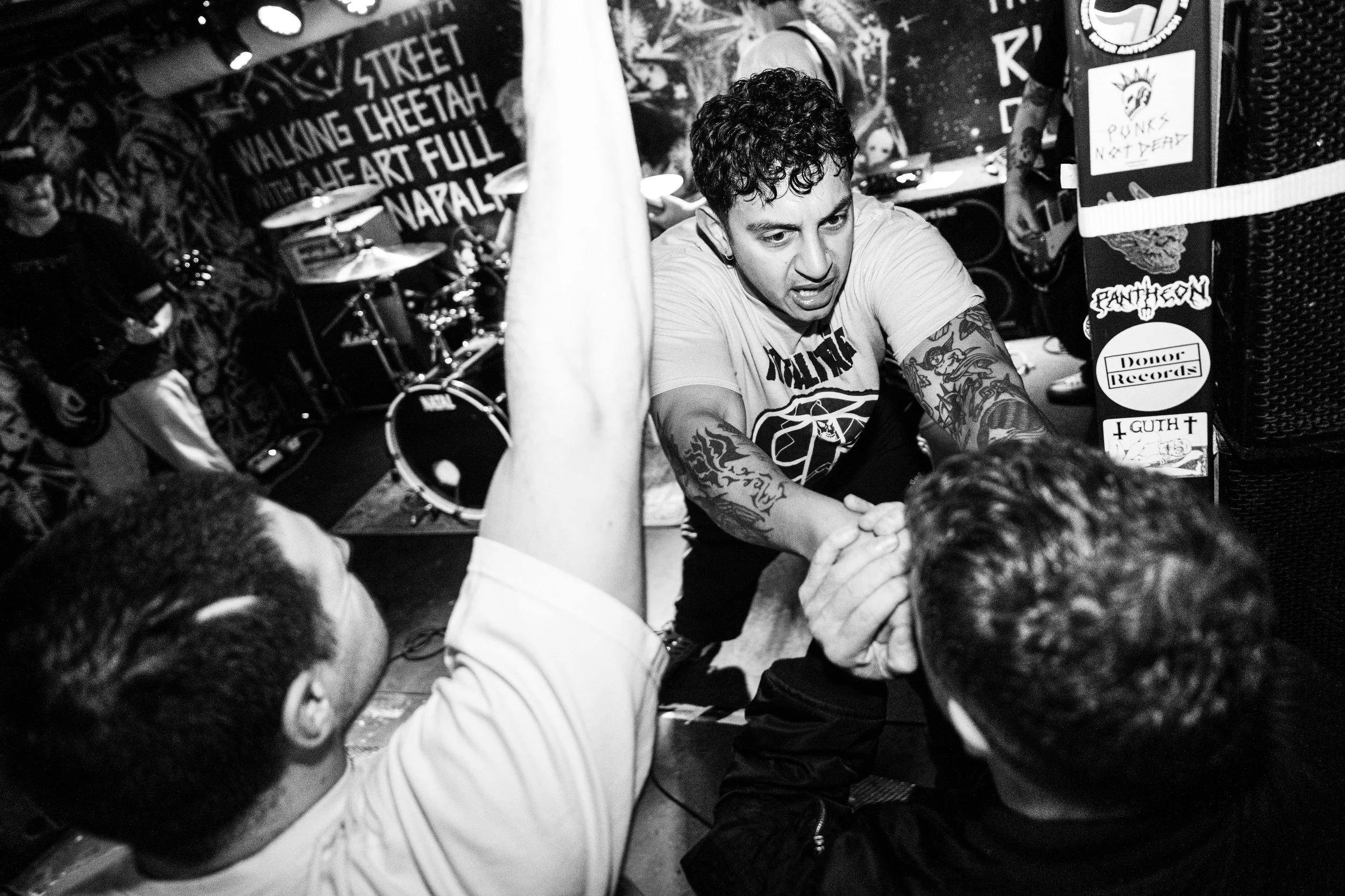 A black and white photo of a lively punk concert showing a man with tattoos on his arms, a graphic t-shirt, and curly hair engaging with fans during an arm wrestling match at a small music venue.