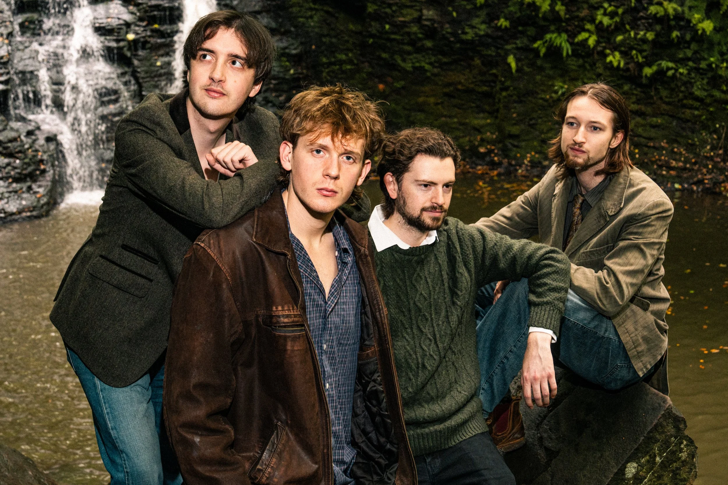 Five young men sitting by a river with a small waterfall in the background, wearing casual and semi-casual clothing, posing for a group photo.