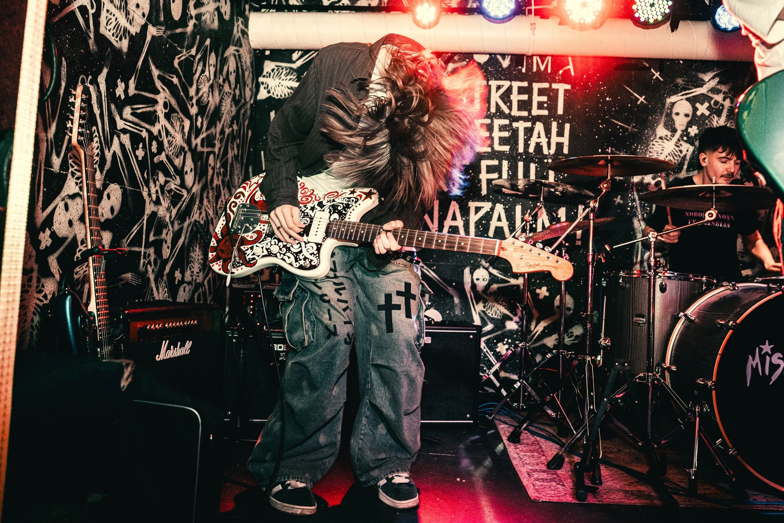 A person with long hair playing an electric guitar on stage, with another person playing drums in the background. The stage has graffiti-style wall art and a sign with city names.