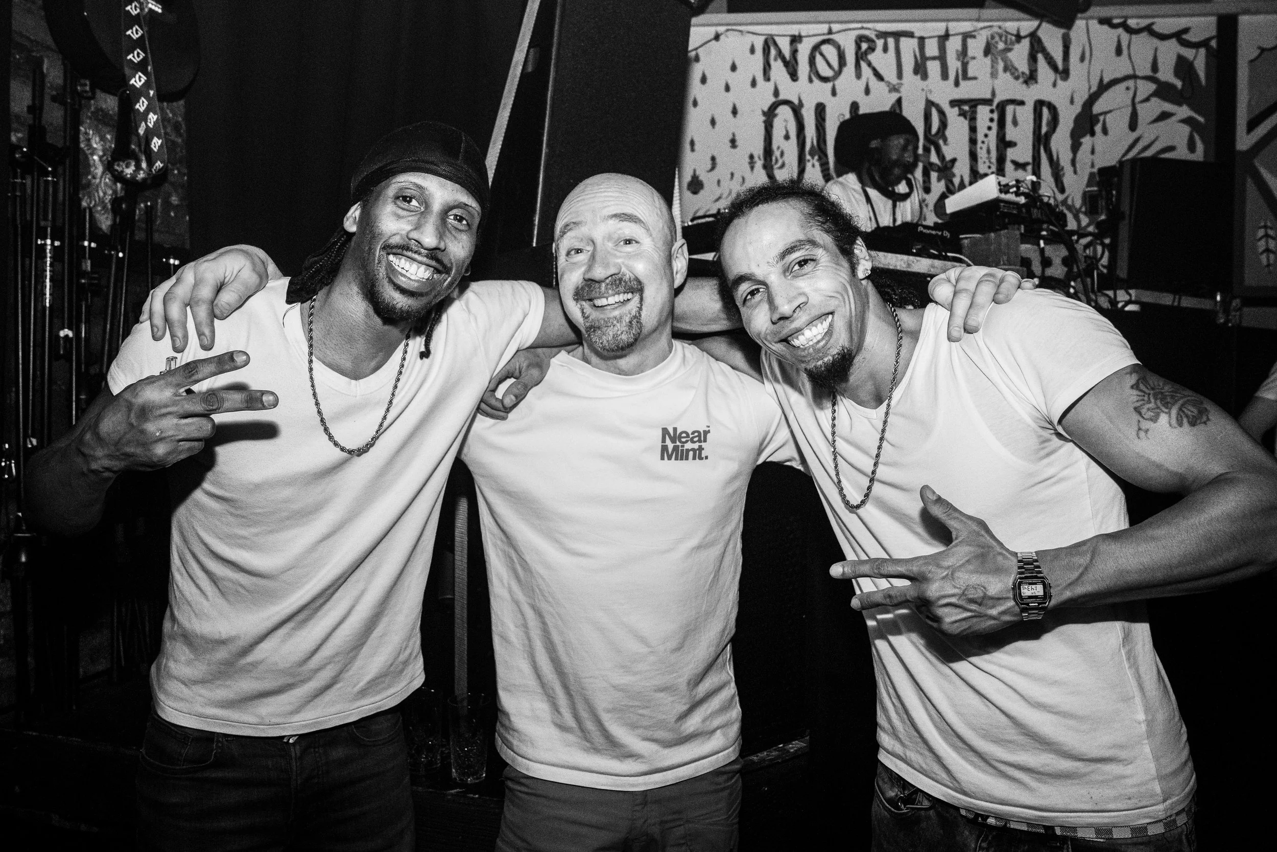 Three men smiling and posing arm-in-arm in a nightclub with DJ equipment and a sign reading 'Northern Quarters' in the background.